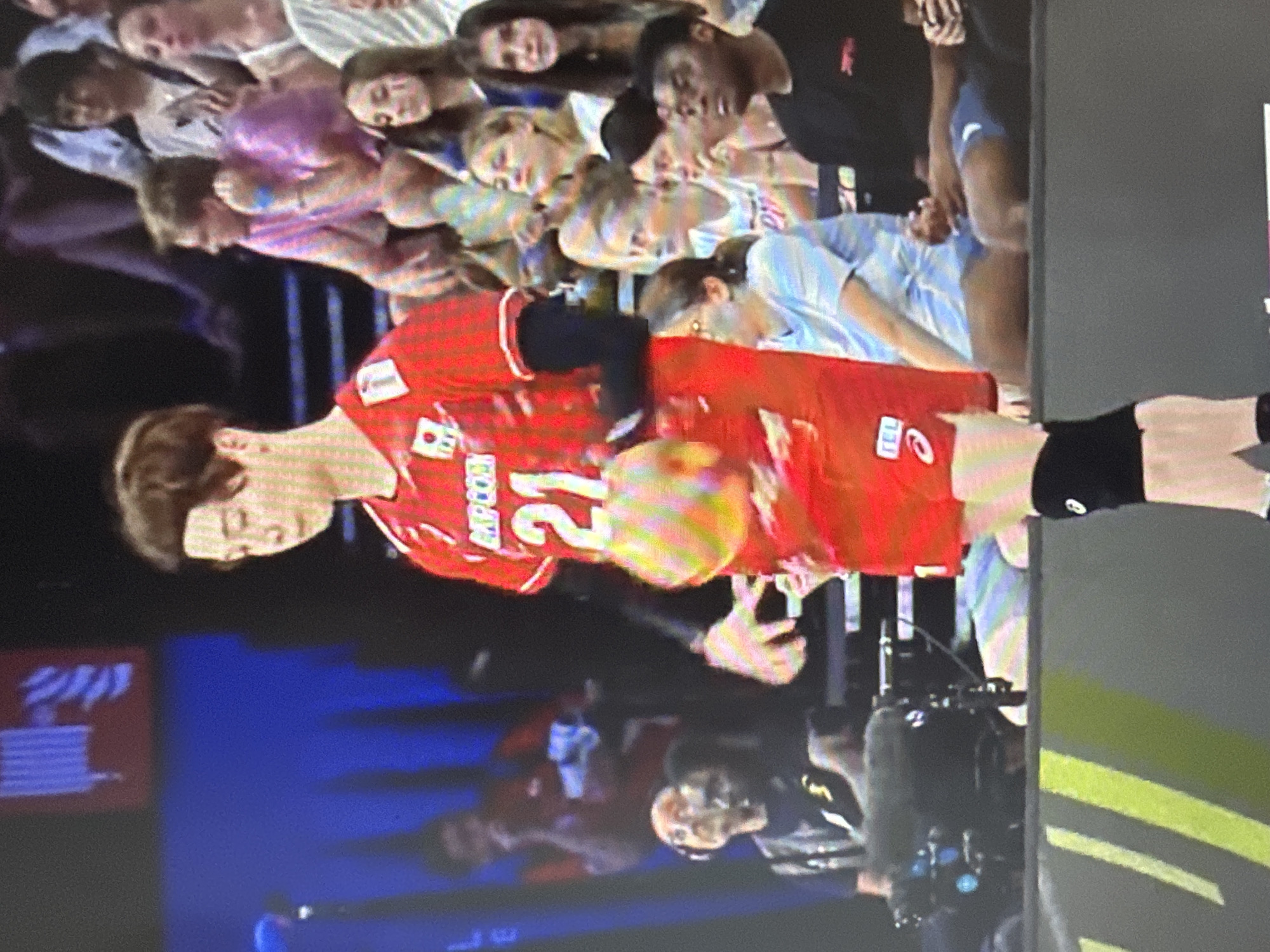 In the media file, a crowd of people watches a volleyball match. The primary subject is a volleyball player in a red uniform with the number 21 on their back, who is in mid-action, potentially serving or receiving. The player is wearing black shorts and arm sleeves. The background shows other players and a seated audience. The scene appears to be indoors, likely a gymnasium, indicated by the lighting and the presence of bleachers. The mood seems to be one of focused attention and possibly excitement from the spectators. Some audience members are looking towards the court, while others are captured in candid moments. The lighting suggests it could be daytime or evening, but there are no clear indicators of weather. Location cues for Peremyshliany, Ukraine, are not visible within the frame. Visible text on the player's uniform includes "RCP COM" and the number "21." There are also some unidentifiable logos on the uniform. The overall image captures a moment of athletic competition and spectator engagement.