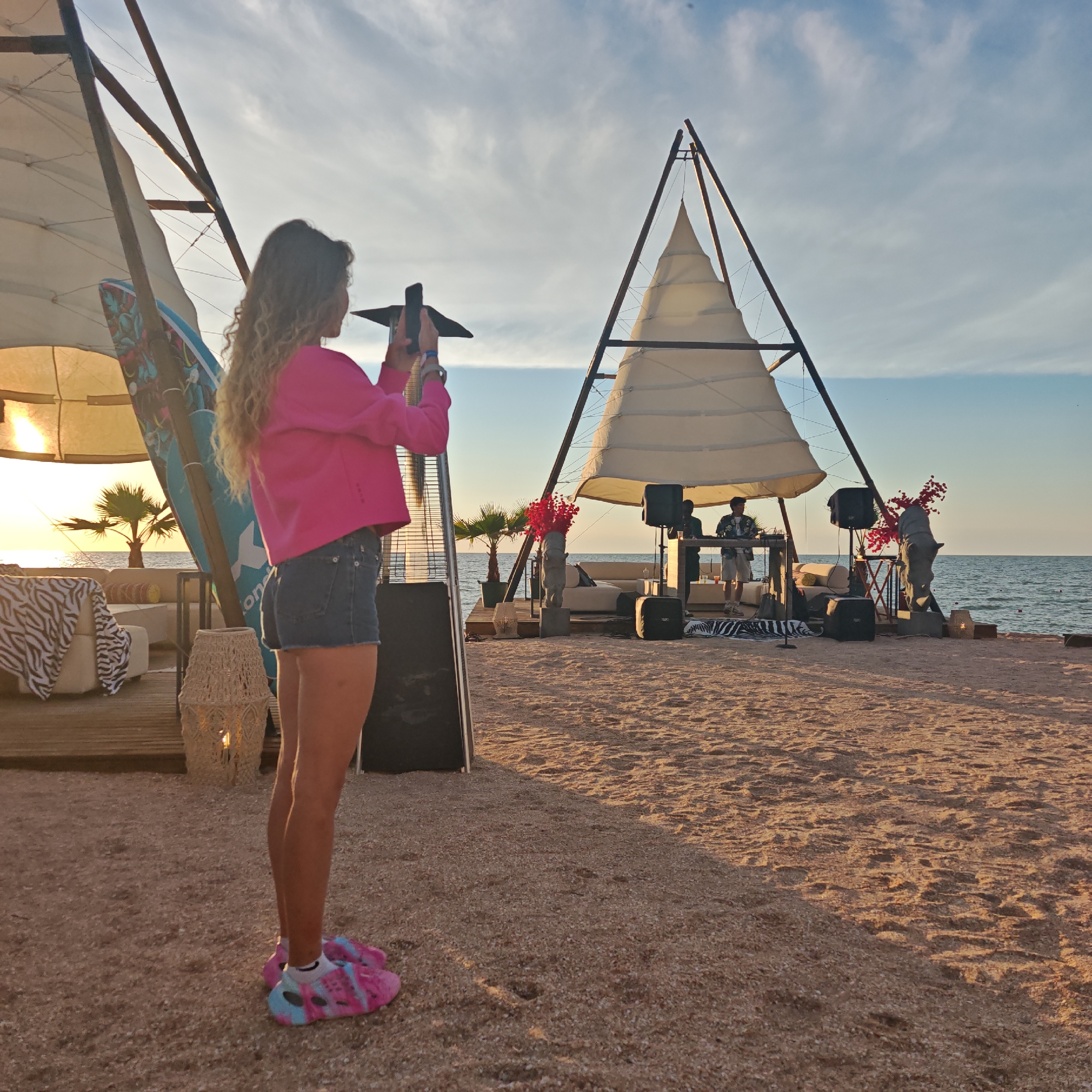 A woman with long, curly blonde hair is standing on a sandy beach, holding a smartphone and taking a picture. She is wearing a bright pink hooded sweatshirt and denim shorts. She has on colorful tie-dye slides. Behind her, a white, sail-like structure is erected on a wooden platform. A DJ booth with two people is set up under the sail. One person is playing music, and the other is observing. Speakers are positioned around the DJ booth. In the background, the calm sea stretches to the horizon under a partially cloudy sky. Palm trees and lounge furniture are visible on the platform, suggesting a relaxed, outdoor event or gathering. The sun is low in the sky, casting a warm glow on the scene, indicating it is either sunrise or sunset. The overall mood appears to be peaceful and enjoyable.