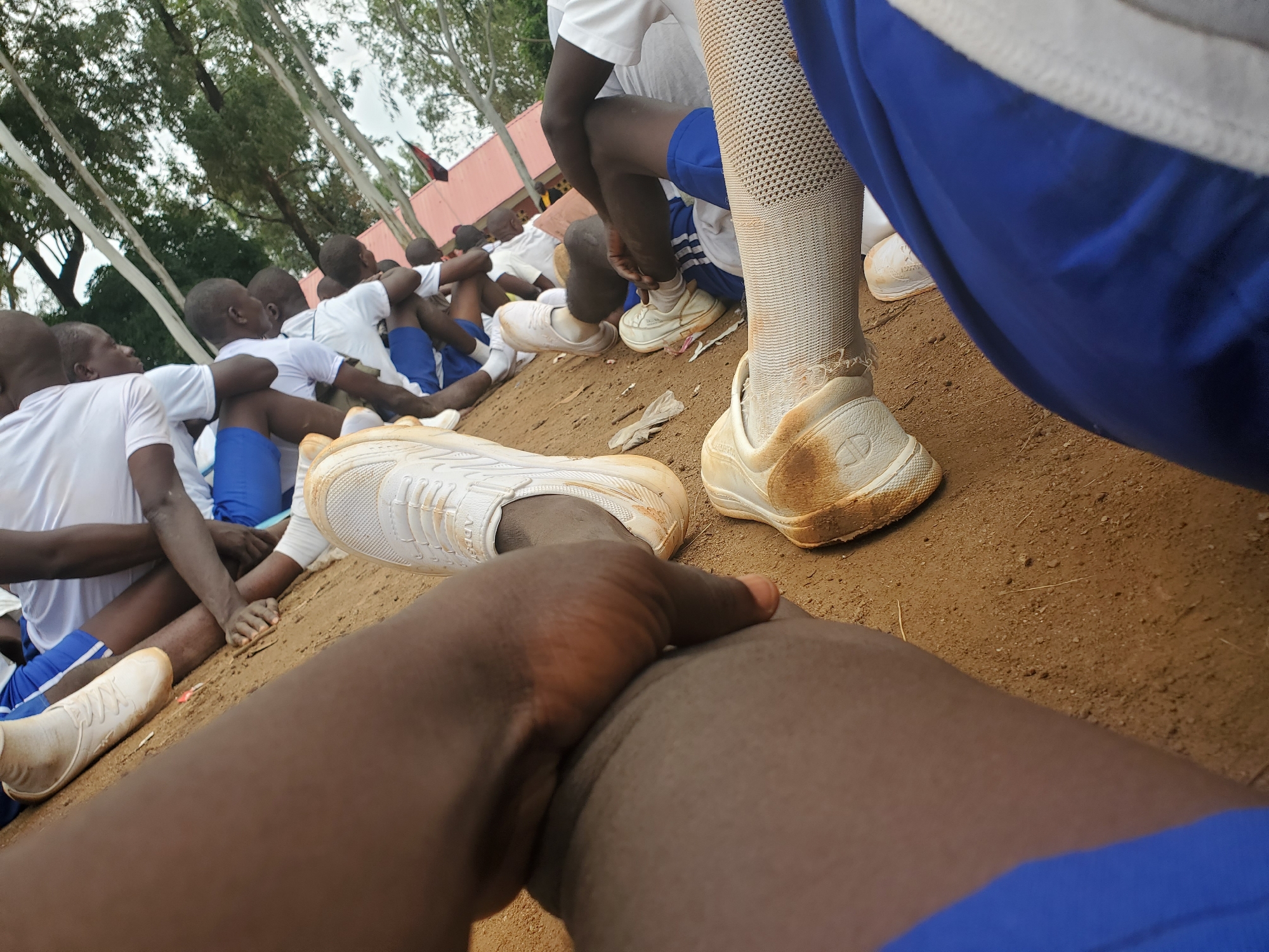 The image depicts a group of young individuals, likely students, gathered outdoors on a dirt surface. They are seated, with some leaning against each other. The individuals are wearing matching white t-shirts and blue shorts, suggesting a school uniform or team attire. Several are wearing white athletic shoes, some of which appear dirty. The environment includes trees and a red-colored building structure in the background, indicating an outdoor setting, possibly a schoolyard or sports facility in Bahit, Nigeria. The scene conveys a sense of rest or waiting.