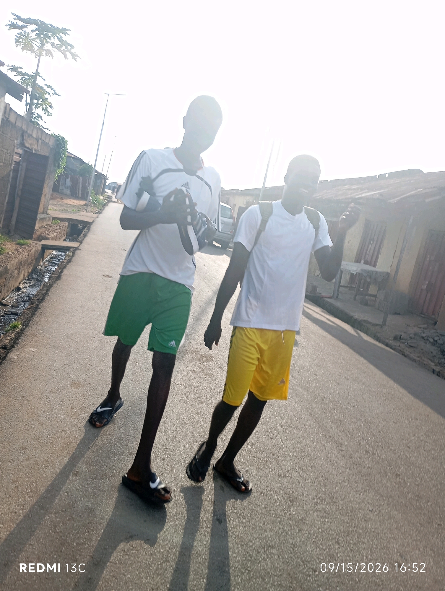 Two young males are walking on an asphalt road in a residential area of Shendam, Nigeria. The male on the left is wearing a white t-shirt and green shorts, holding a pair of black and white athletic shoes. The male on the right is wearing a white t-shirt and yellow shorts, with a backpack slung over his shoulders. Both are wearing black flip-flops. The road is flanked by buildings with corrugated metal roofs and a drainage ditch on the left. The sky is bright and overcast, casting long shadows on the road. The timestamp "09/15/2026 16:52" is visible in the lower right corner, and "REDMI 13C" is visible in the lower left corner.