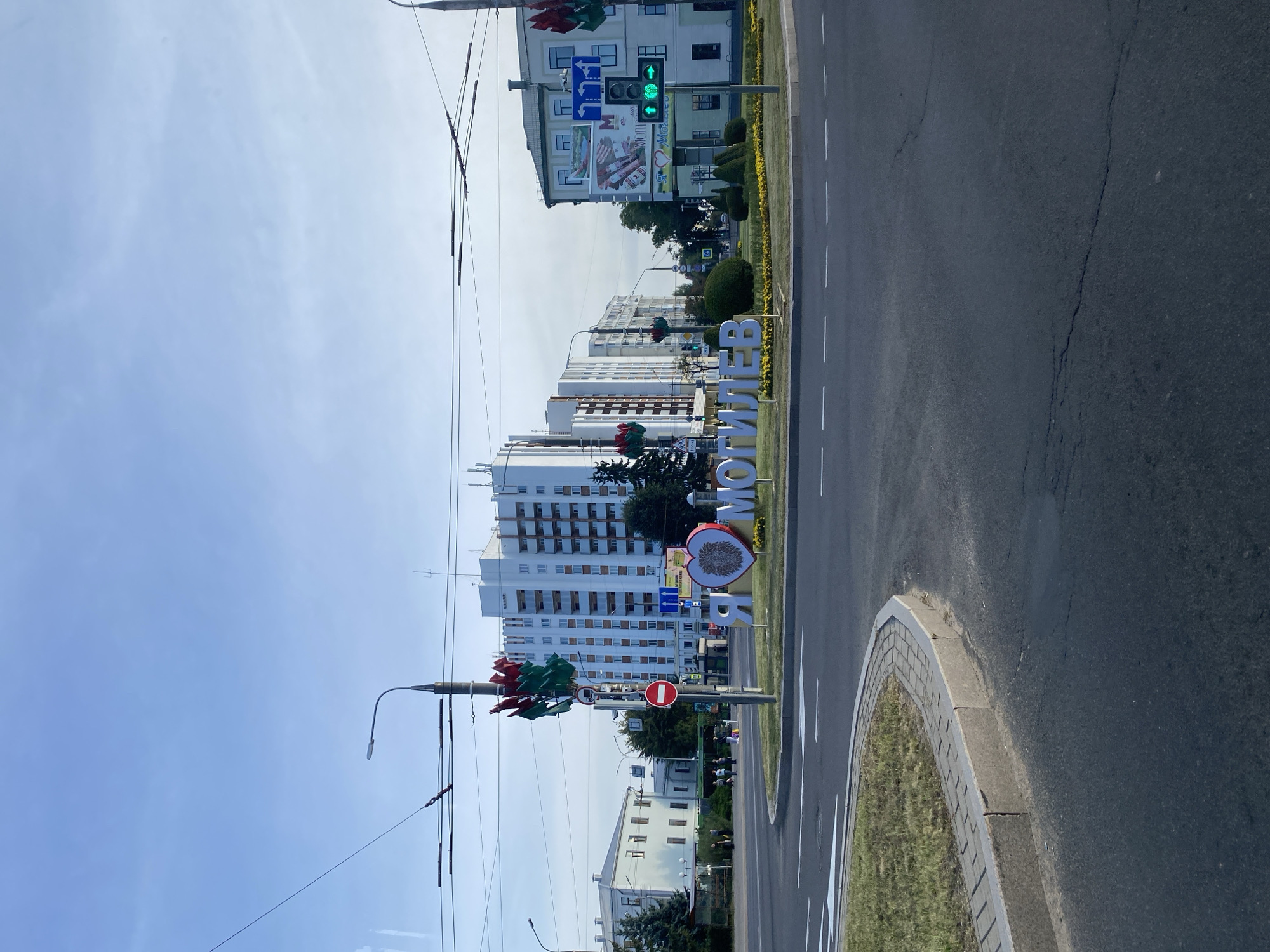The image captures an outdoor urban scene in Mogilev, Belarus. In the foreground, a paved road with white lane markings curves around a grassy roundabout. A concrete curb separates the road from the roundabout. Dominating the center of the roundabout is a large, three-dimensional art installation spelling out "Я ЛЮБЛЮ МОГИЛЕВ" (I Love Mogilev). The word "ЛЮБЛЮ" is depicted as a heart shape with a stylized lion's head design in its center. Behind the art installation, various buildings are visible. To the right, a multi-story building with a light-colored facade and numerous windows stands. A traffic light with a green arrow is illuminated. Further back, another white multi-story building with a more modern architectural style is prominent. To the left, a smaller, older-looking building with a light facade and arched windows can be seen, along with some trees and a bus stop structure. Overhead, electrical wires and street lights crisscross the sky, which is a clear light blue. There are no people clearly visible in the foreground or engaging in any specific actions. The overall impression is that of a public space within a city.