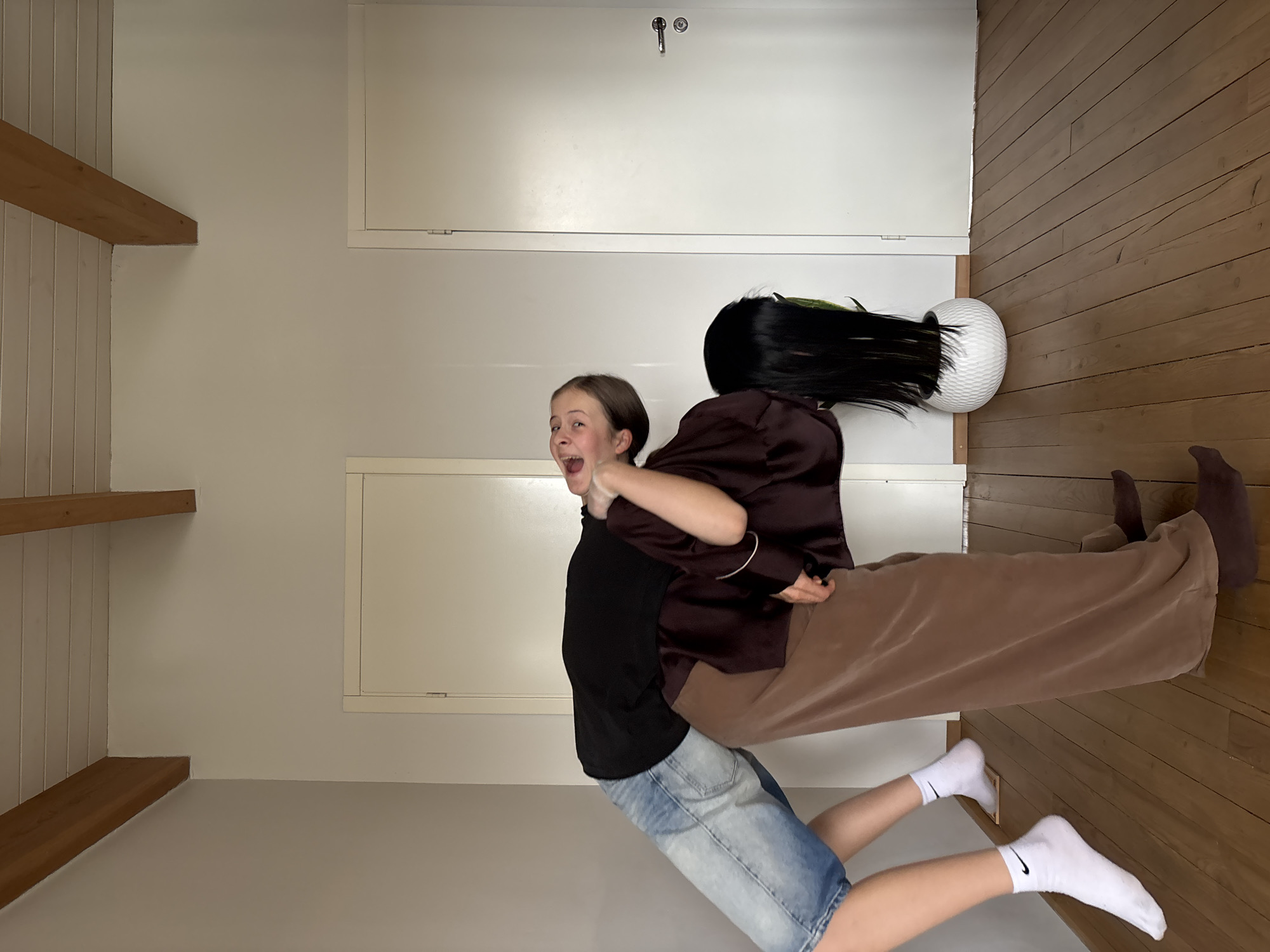 The image, set in Chaikovsky, Russia, captures a humorous indoor scene featuring two individuals engaged in a playful interaction, enhanced by a visual illusion. The photograph is oriented with a light wooden floor running along the right side and light-colored walls, featuring wooden shelves and panels, on the left.
In the foreground, a young person with light brown hair, wearing a black t-shirt and blue denim shorts, reacts with a wide-open mouth, conveying surprise, laughter, or excitement. Their arms are raised, seemingly interacting with the other person. They wear white socks prominently displaying a black Nike swoosh logo.
To their right, a second individual's head is replaced by an illusion: a white, textured spherical object, similar to a planter, holds a long, straight black wig, which appears to have green highlights at the top. This person wears a dark brown or burgundy satin-like jacket and wide-leg brown pants.
The background shows a simple, brightly lit room with white walls, two wooden shelves on the left, and a white door or cabinet with a metal handle on the far wall. The overall mood is lighthearted and suggests a staged, amusing moment or a visual prank.