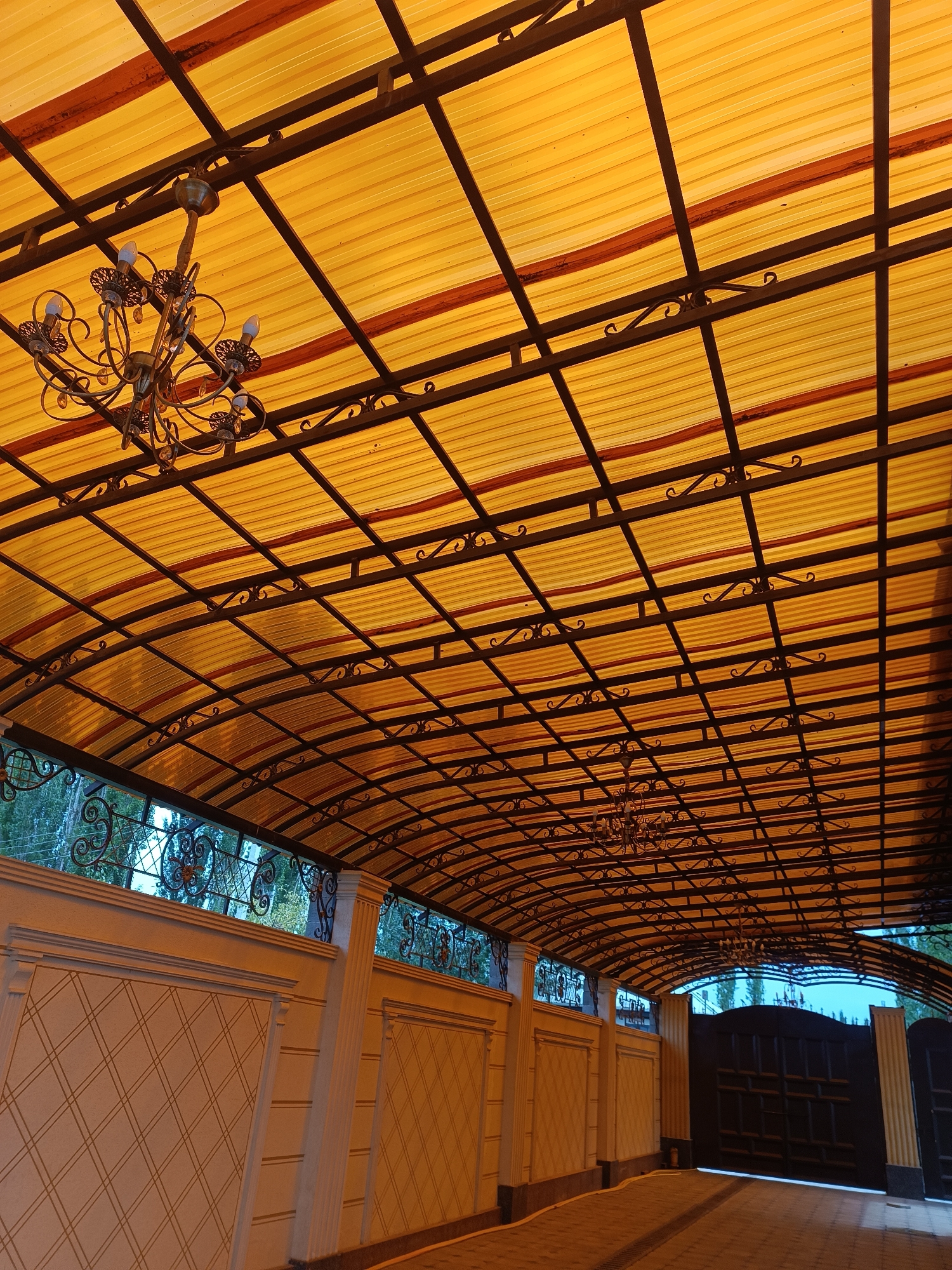This image depicts a covered walkway with a yellow, ribbed translucent roof supported by a dark metal frame. Ornate chandeliers hang from the ceiling. The walkway is bordered by decorative cream-colored walls with diamond-patterned panels. In the background, a large, dark metal gate is visible, suggesting an entrance or exit. The scene is set in Myrza-Ake, Kyrgyzstan, characterized by its architectural style and materials. The lighting indicates it may be dusk or dawn. No people are present in the image.