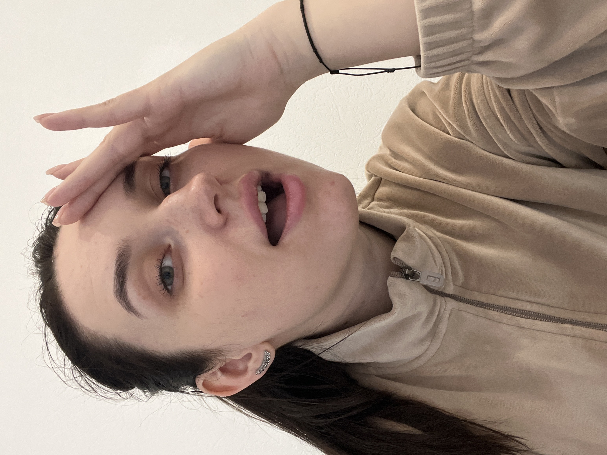 A woman with dark hair is pictured in a close-up selfie, with her head tilted. She is wearing a beige velour zip-up jacket. Her right hand is raised to her forehead. She has light skin, blue eyes, and is wearing a small stud earring. Her mouth is open. A thin black string bracelet is visible on her wrist. The background is a plain white wall. There is no visible indication of the city of Tyumen, Russia, or any other specific location.