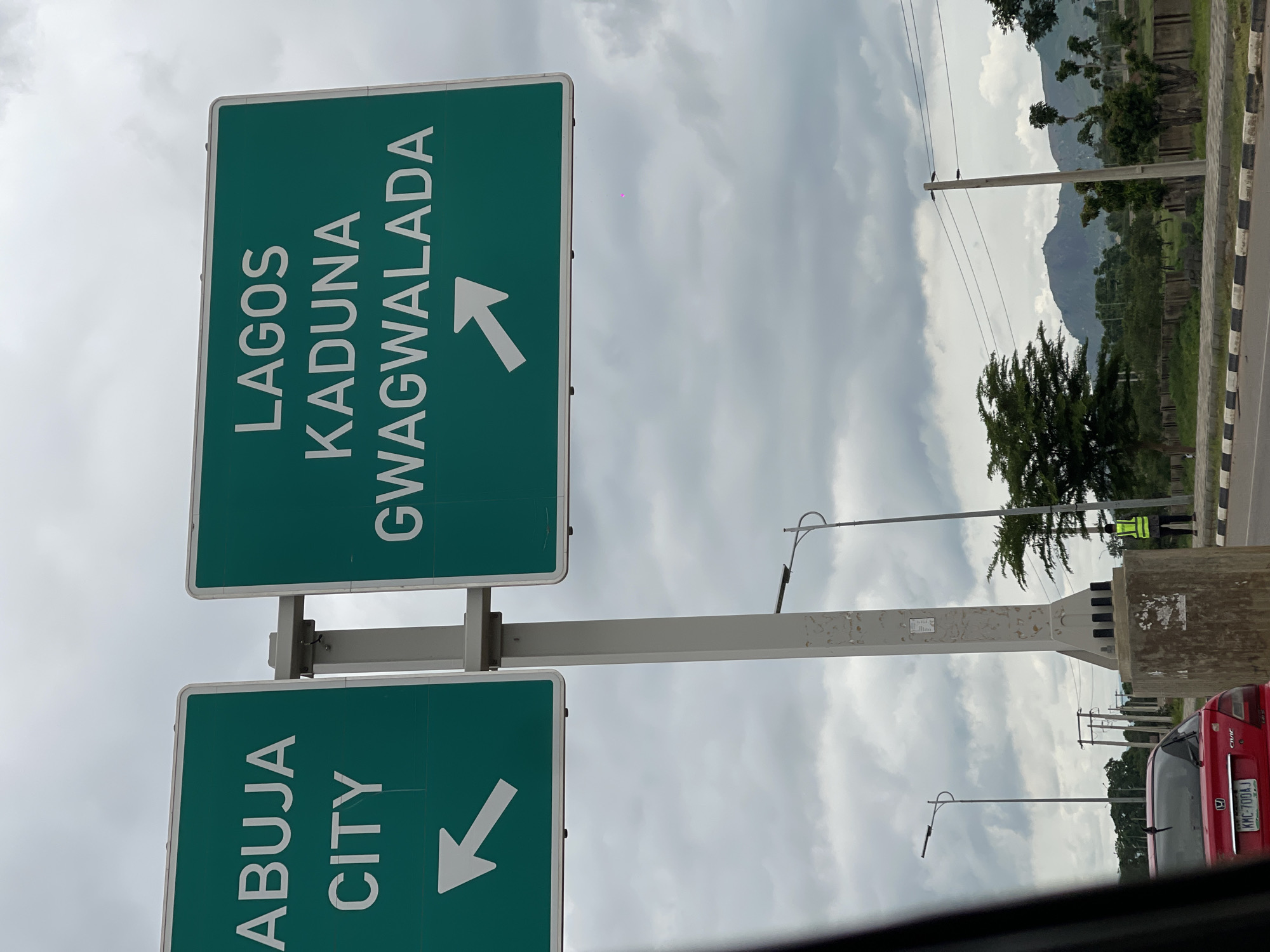 An eye-level, medium outdoor shot features two green directional highway signs against a cloudy sky. The sign on the left reads "ABUJA CITY" in white capital letters and has a white arrow pointing left. The sign on the right reads "LAGOS KADUNA GWAGWALADA" in white capital letters and has a white arrow pointing right. Both signs are mounted on a gray pole. In the background, a red car is parked on the right side of the road. A person in a yellow safety vest stands on the sidewalk. The surrounding environment includes trees, mountains in the distance, and power lines. The scene appears to be located near Immigration Quarters, Nigeria.