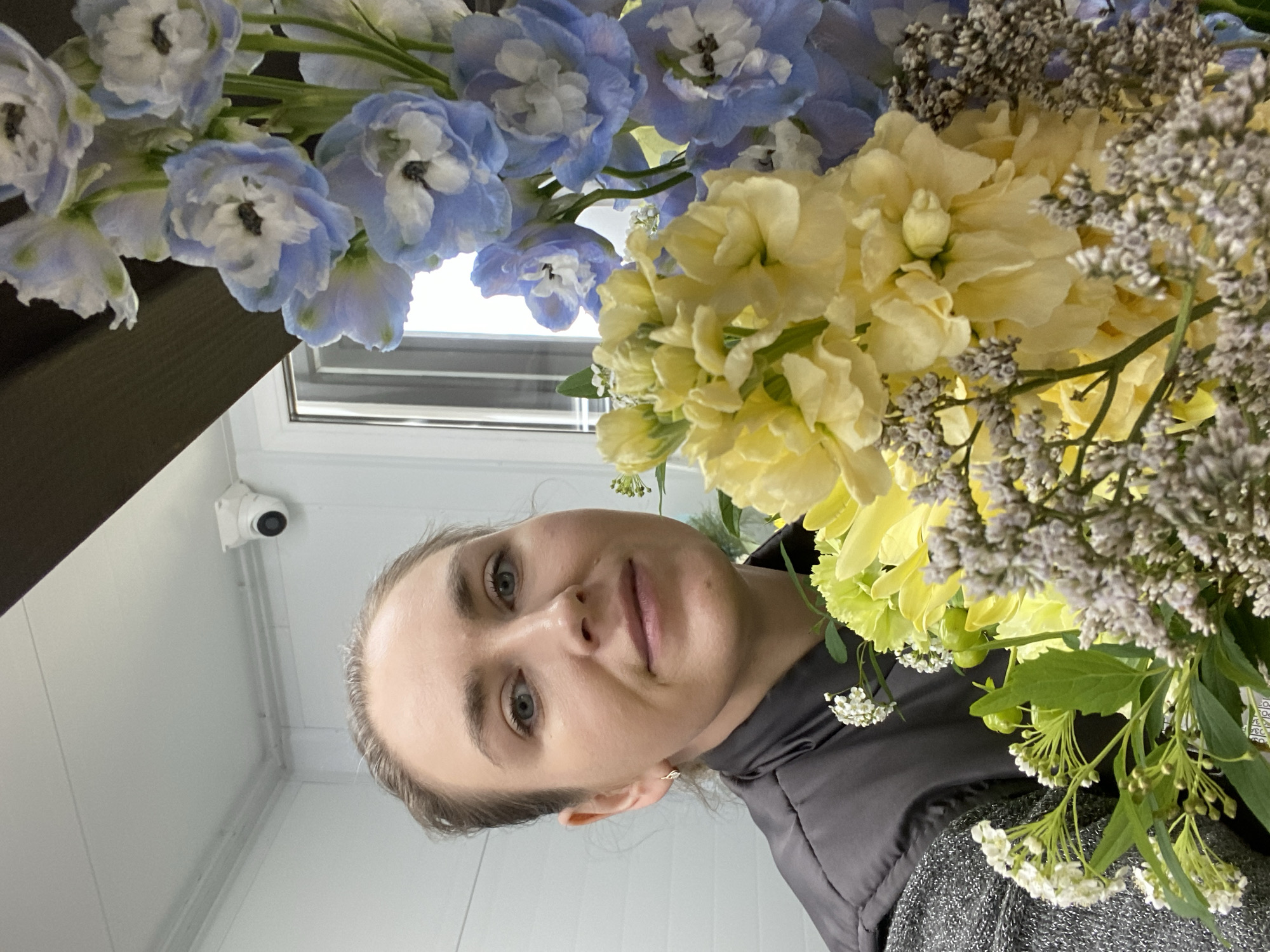 A woman is positioned in the foreground, partially obscured by a bouquet of flowers. The flowers are a mix of blue and white delphiniums, yellow blooms, and smaller white flowering branches. The woman's face is visible, featuring fair skin, light eyes, and her hair is pulled back. She wears a dark gray jacket with a textured sweater underneath. The background features a white interior wall and a window with a view of the sky. A security camera is visible on the ceiling. There is no location information within the frame.