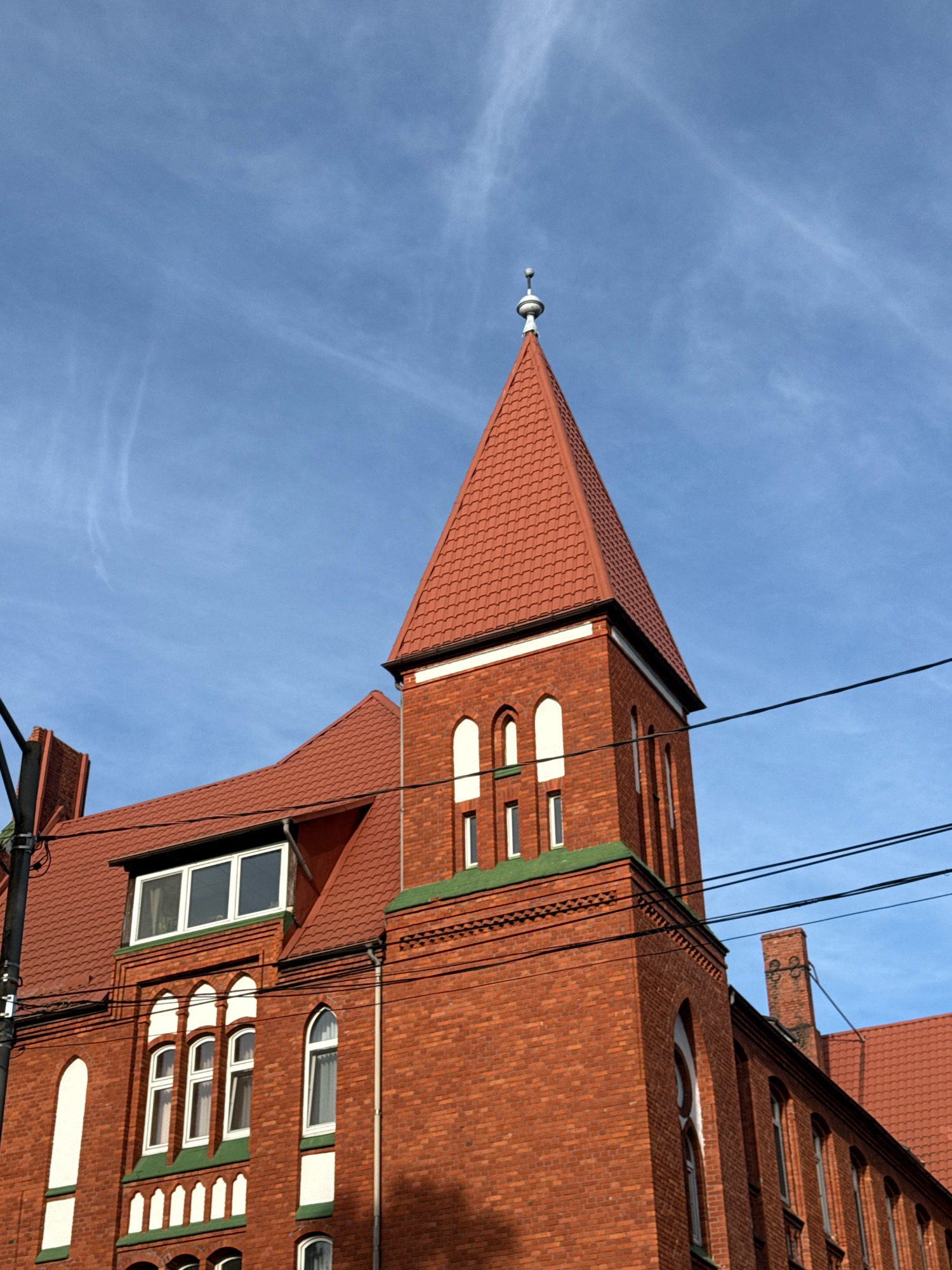 The image shows a close-up, low-angle shot of the brick facade of a building with a red-tiled roof and a prominent tower, against a bright blue sky with faint white clouds. The building appears to be architecturally significant, with arched windows and decorative brickwork. The scene is situated in Kaliningrad, Russia, specifically depicting the Saint Nikolai Orthodox Church. The building's architecture suggests a historical context, possibly dating back to the late 19th or early 20th century. There are no people visible in the frame. Overhead, electrical wires cross the sky in front of the building. The lighting suggests it is daytime, with the sun casting shadows on the brickwork.