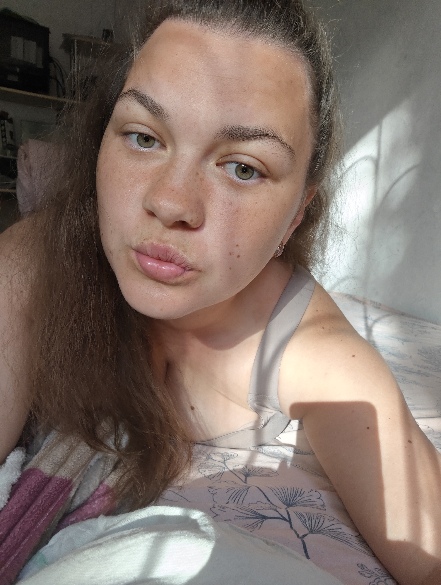 A fair-skinned woman with freckles and light brown hair is lying in bed. She is wearing a gray tank top and earrings. The woman is making a kissing face. Sunlight is filtering into the room and casting shadows on the wall behind her and on her right arm. Bedding with a floral pattern is visible underneath her. A striped blanket is partially visible on the left side of the frame. A shelf with objects is slightly out of focus in the background. The location is assumed to be a residential interior, although the city of Chaikovsky, Russia cannot be independently verified.
