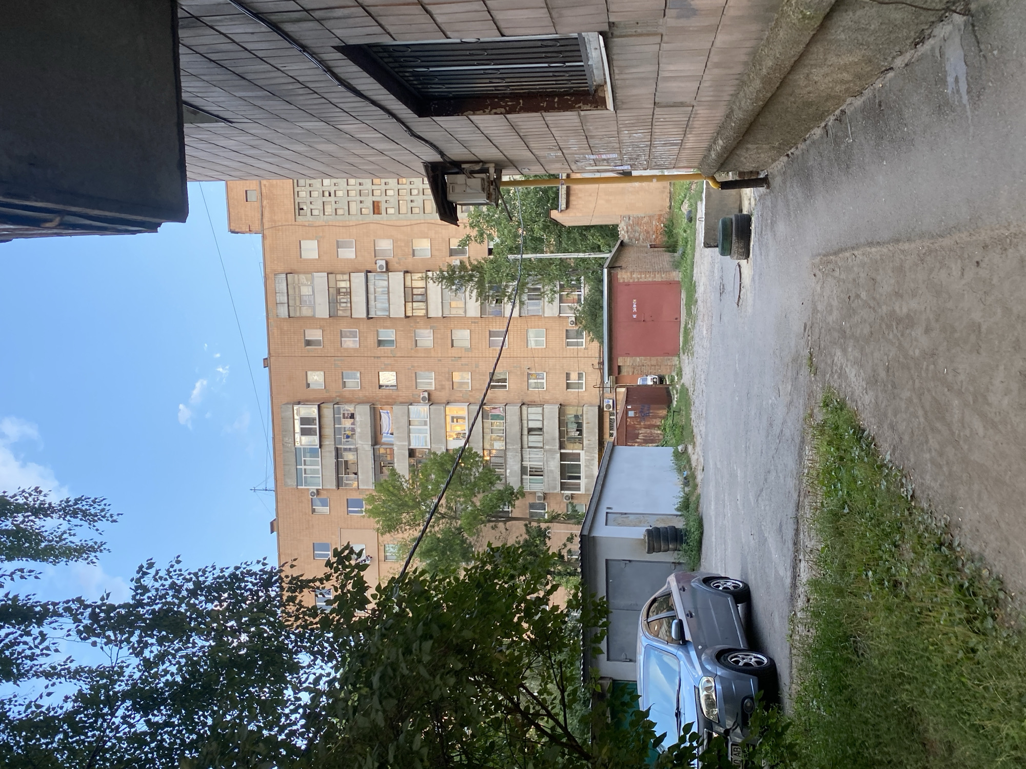 The image depicts a residential area in Poltava, Ukraine, likely a courtyard. In the foreground, a grey sedan is parked on an asphalt surface. To the left, lush green grass and foliage are visible. Adjacent to the car, a light blue corrugated metal garage with grey doors stands, with a stack of black tires to its right. Further back, a red corrugated metal garage with the Cyrillic text "НЕ ЗАБИРАТИ" (Do not take) is visible. Behind these garages, a large, multi-story brick apartment building with numerous windows and balconies dominates the background. A tall, slender green tree is situated in front of the building. To the right of the frame, a section of a brick building with a barred window is present. A yellow utility pole with a small grey box attached to it is also visible. The sky above is a clear blue with a few wispy white clouds. The scene appears to be set during daylight hours.