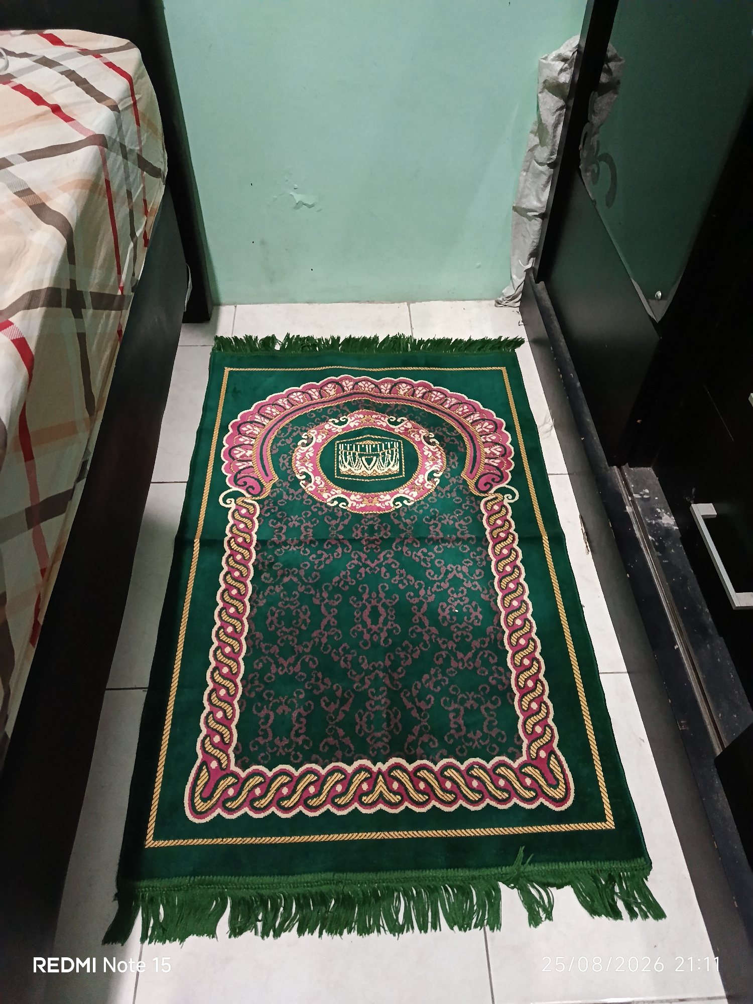 A prayer rug lies on a white tiled floor in a room. The rug is primarily green with intricate patterns in shades of pink and gold. It features fringed edges at both ends and a decorative border. To the left of the rug is the side of a bed with a striped comforter in cream, brown, and red. To the right of the rug is a dark wooden piece of furniture, possibly a wardrobe or cabinet, partially covered by a light gray plastic sheet. The wall behind the rug is painted a light blue-green color. The overall scene appears to be in a domestic setting.
