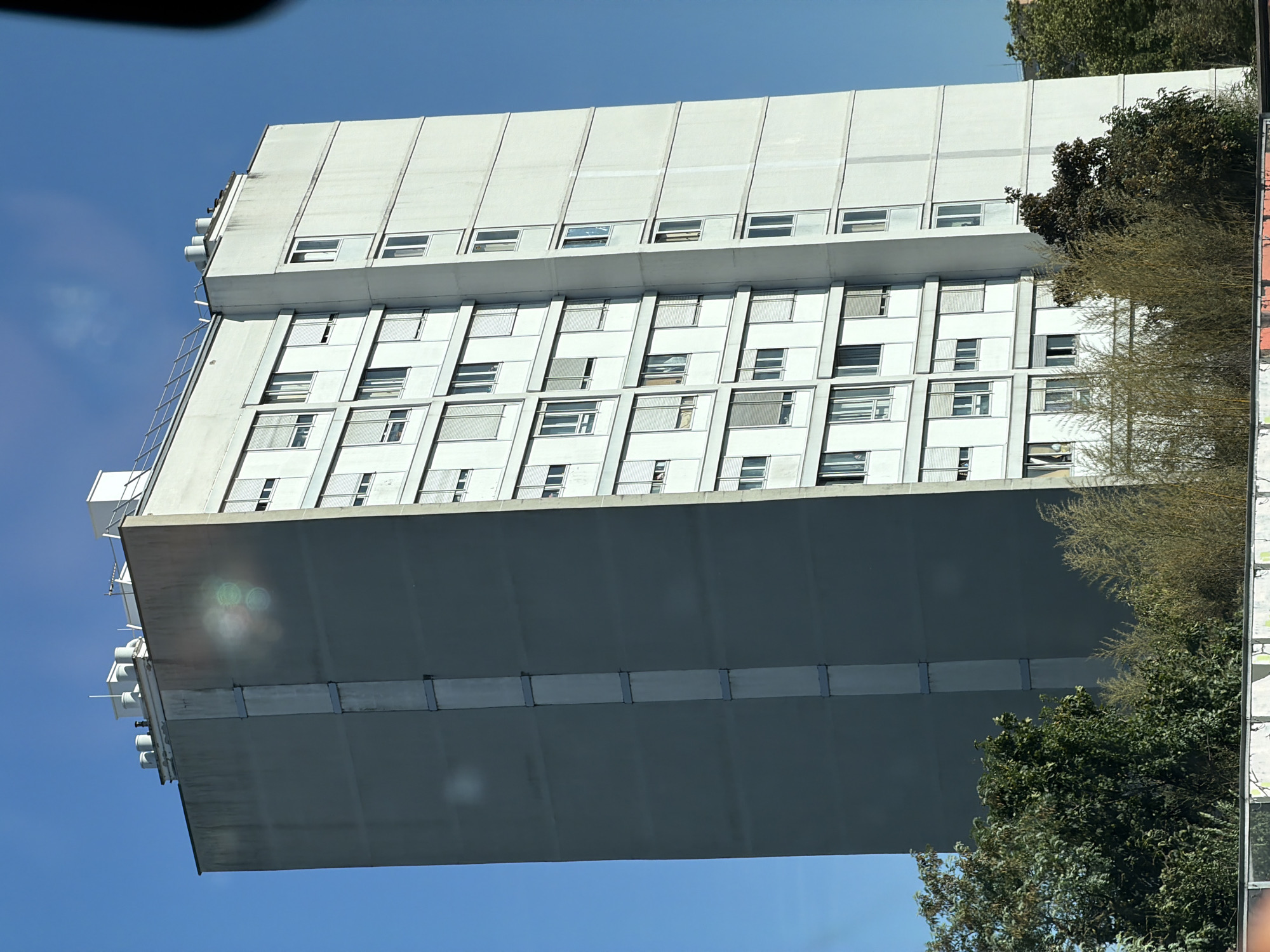 An exterior, medium-range, eye-level shot captures a tall, grey residential apartment building in Bron, France. The building features a light-colored facade on its right side, punctuated by numerous windows, many equipped with white shutters. The left side of the building is a uniform grey with a vertical band of metal fixtures running its height. The top of the building is flat and includes several ventilation units and what appears to be a solar panel array. The sky is a clear, bright blue, with some lens flare visible on the grey section of the building. Trees and foliage are present at the base of the building, obscuring the lower levels. The overall impression is of a functional, urban residential structure under a sunny sky.
