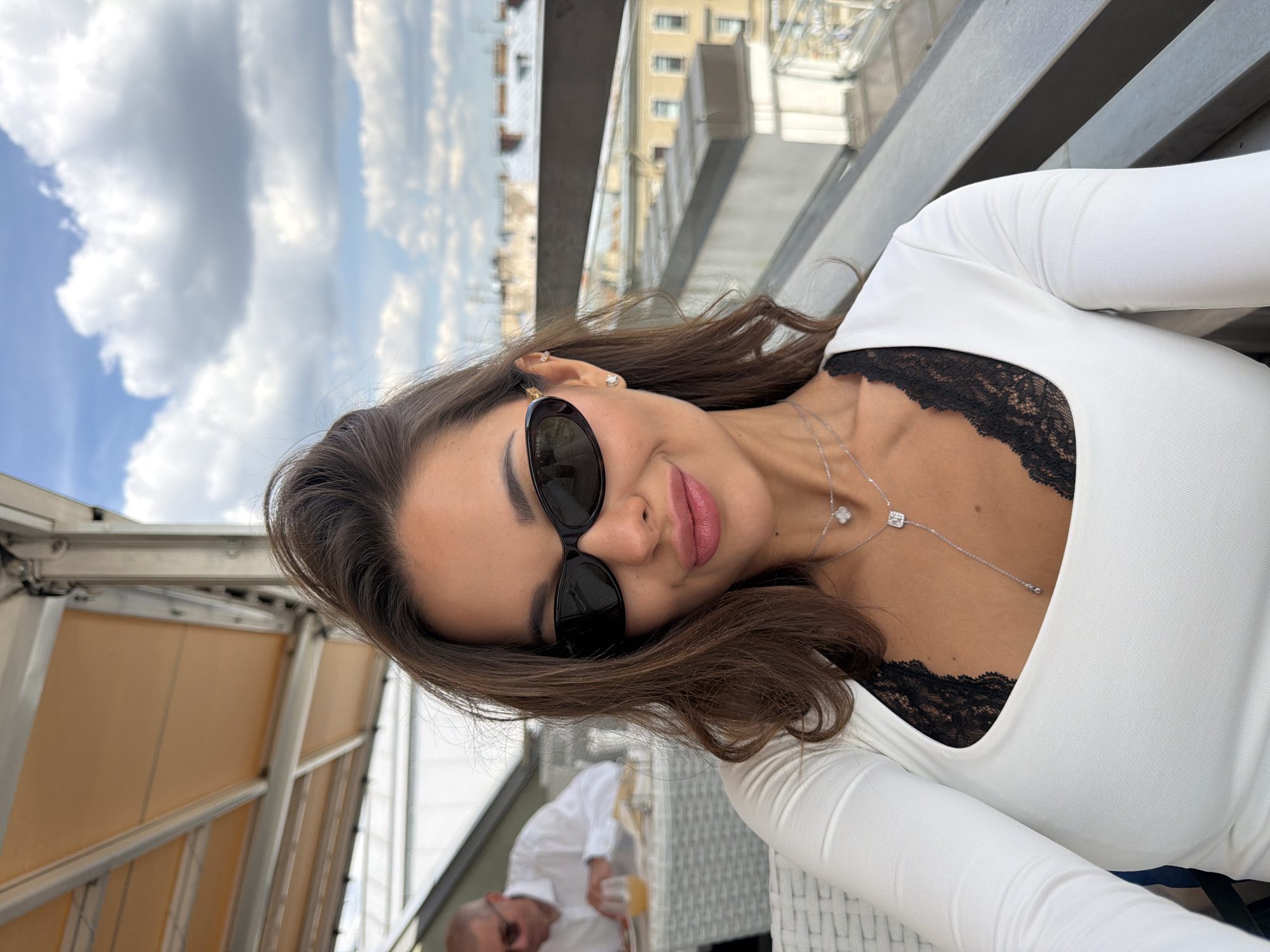 A woman with long, dark brown hair and sunglasses is taking a selfie from a rooftop restaurant in Saint Petersburg, Russia. She is wearing a white, long-sleeved top with a black lace trim visible at the neckline. She also has a delicate silver necklace with a pendant. In the blurred background, another person in a white shirt is seated at a table, and a cityscape with buildings and a cloudy sky is visible. The setting appears to be an outdoor dining area with a railing.