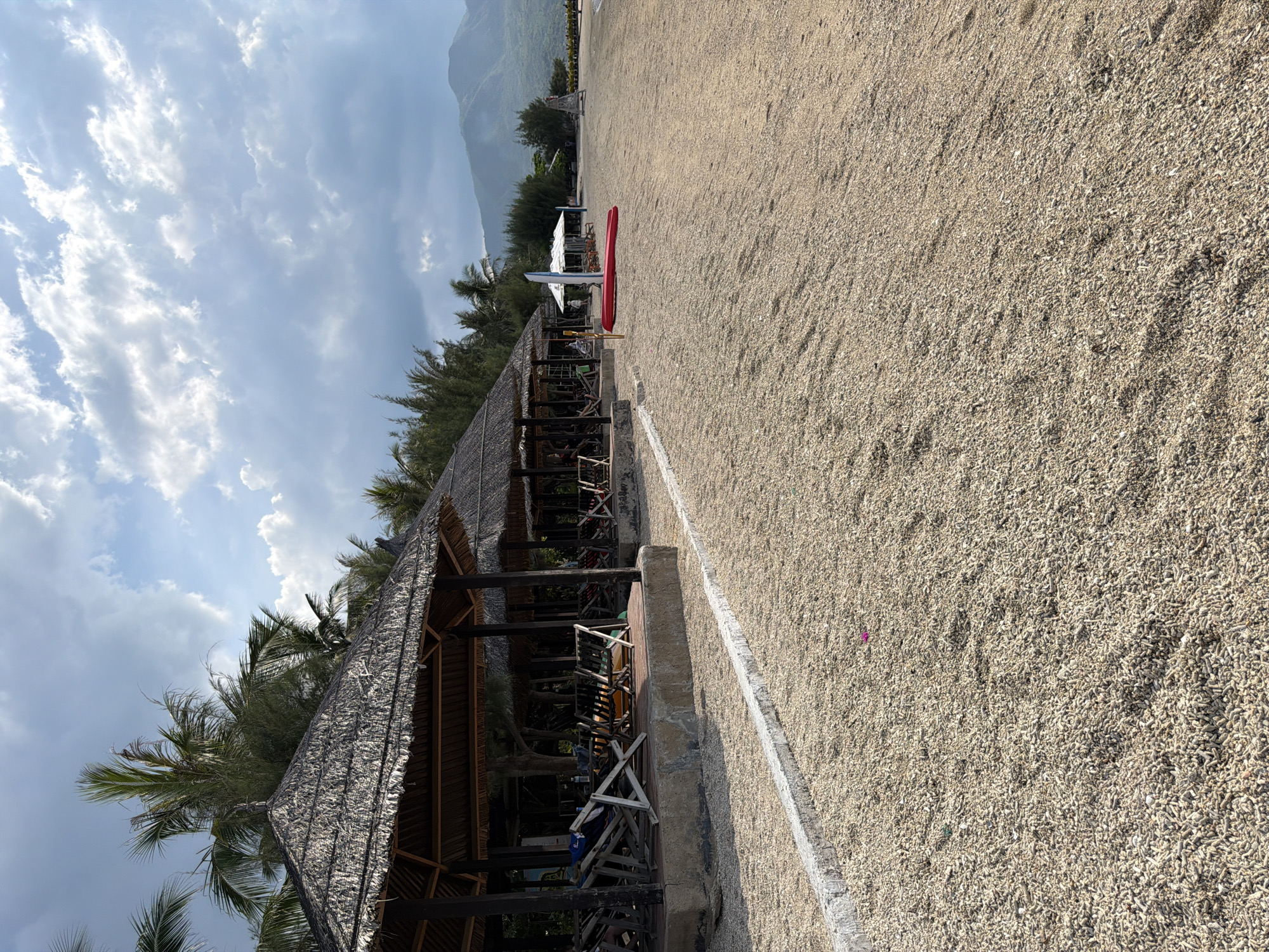 The image depicts a tranquil beach scene in Ninh Ích, Vietnam. Several thatched-roof cabanas line the shore, providing shade for rows of lounge chairs. The beach itself is composed of small, light-colored pebbles and shells. In the distance, lush green mountains rise against a sky filled with dramatic, grey-tinged clouds. Two red kayaks are visible on the sand near a white sail, suggesting recreational activities. The overall impression is one of a peaceful, natural setting, possibly a resort area.