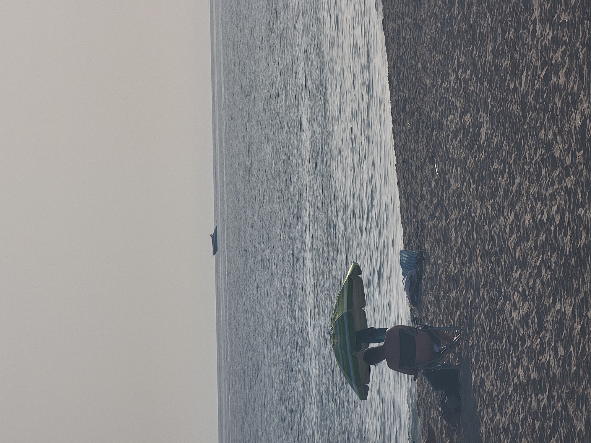 The image depicts a serene beach scene in Quarteira, Portugal, with the vast Atlantic Ocean stretching towards the horizon under a pale, overcast sky. In the foreground, coarse sand forms the beach, marked by ripples from receding waves. A solitary figure is seated in a folding chair, partially shaded by a green and white striped beach umbrella. The individual is facing away from the camera, towards the sea, with their back visible. Beside the chair, beach bags and a towel are placed on the sand. Further out at sea, a sleek, dark-colored boat is visible, cutting through the calm waters. The overall atmosphere is tranquil and peaceful, suggesting a quiet moment of relaxation by the coast.