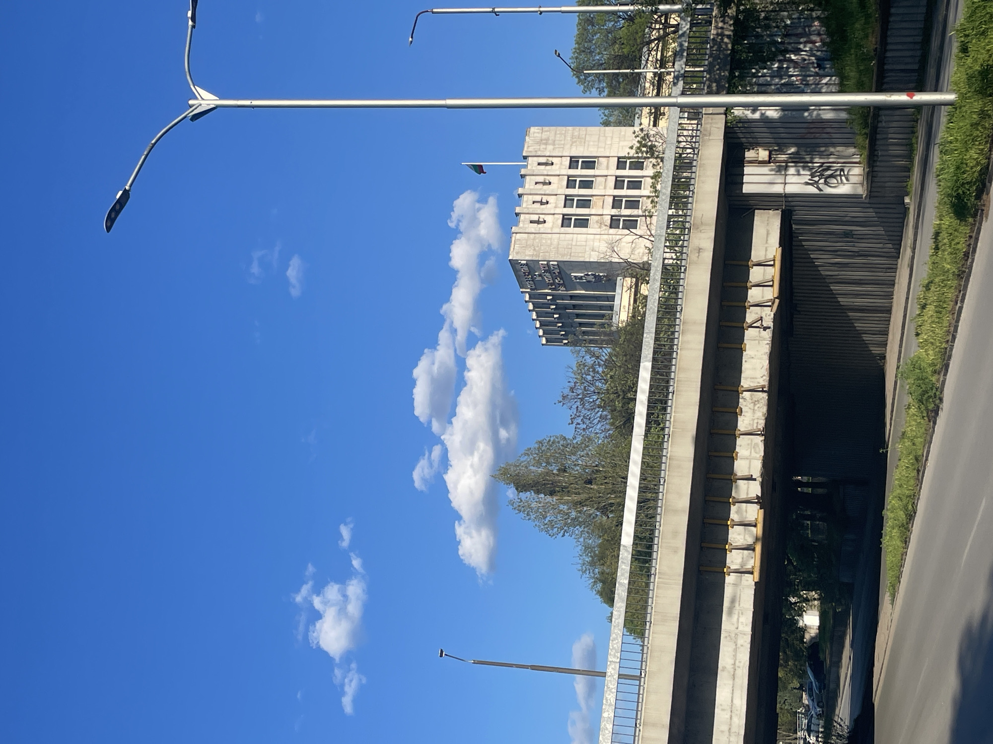 This image captures a static, sunny urban scene in Plovdiv, Bulgaria, focusing on infrastructure and architecture under a clear blue sky.
The primary subjects are a concrete overpass or bridge and a building in the background. The overpass features sturdy concrete construction with metal railings running along its length. Several modern streetlights, each with multiple luminaires, extend from tall poles, arcing over the scene. Beneath the overpass, yellow support beams are visible, suggesting structural work or reinforcement. A road or pathway passes underneath, bordered by green grass and another paved area to the right.
In the midground, a prominent, multi-story building stands with a concrete facade and numerous rectangular windows. A Bulgarian flag, identifiable by its horizontal white, green, and red stripes, flies from a flagpole on the roof of this building.
The weather is bright and sunny, indicated by the vivid blue sky dotted with scattered white cumulus clouds. The time of day appears to be midday or early afternoon, with ample natural light. There are no people or specific activities taking place, presenting a quiet moment in the city.
A notable detail is the graffiti on the concrete wall of the underpass structure, to the right side of the image, which appears to be a black tag, possibly spelling "LEON" or a similar word. The Bulgarian flag serves as a strong location cue, confirming the context of Plovdiv.
