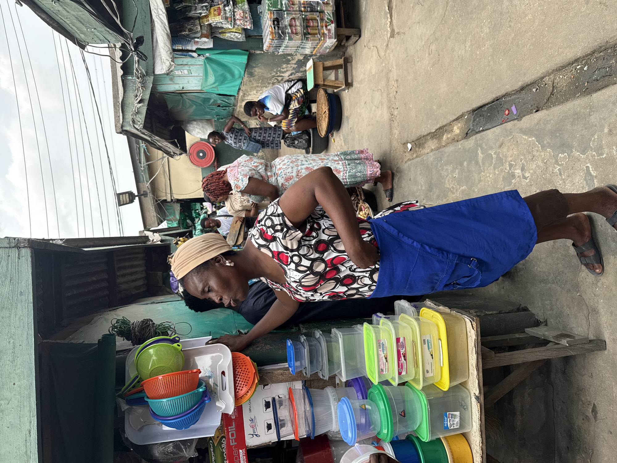 This image captures a vibrant street market scene in Lekki, Nigeria, under an overcast sky. The primary subject is a woman in the foreground, wearing a patterned top with red and white circles, a blue skirt, and a light-colored head wrap. She is leaning over a stall, appearing to examine items for sale. Her left arm rests on the stall, which is laden with various household goods, including colorful plastic colanders, bowls, and multiple clear plastic food storage containers with colored lids (green, blue, yellow, purple). A box with "YAM LIFE Premium FOIL" printed on it is also visible on the stall.
In the background, several other individuals, mostly women, are engaged in market activities. One woman, wearing a light-colored floral dress, stands further back, while another woman sits on a low stool beside what appears to be a large woven basket or tray, with packaged goods displayed on shelves behind her. Overhead, numerous power lines crisscross against the sky, and various market stalls constructed from simple materials like corrugated metal and tarpaulins line the narrow path. The ground is concrete, showing signs of wear and tear, with a visible drainage channel. The scene depicts a bustling, everyday commercial environment.