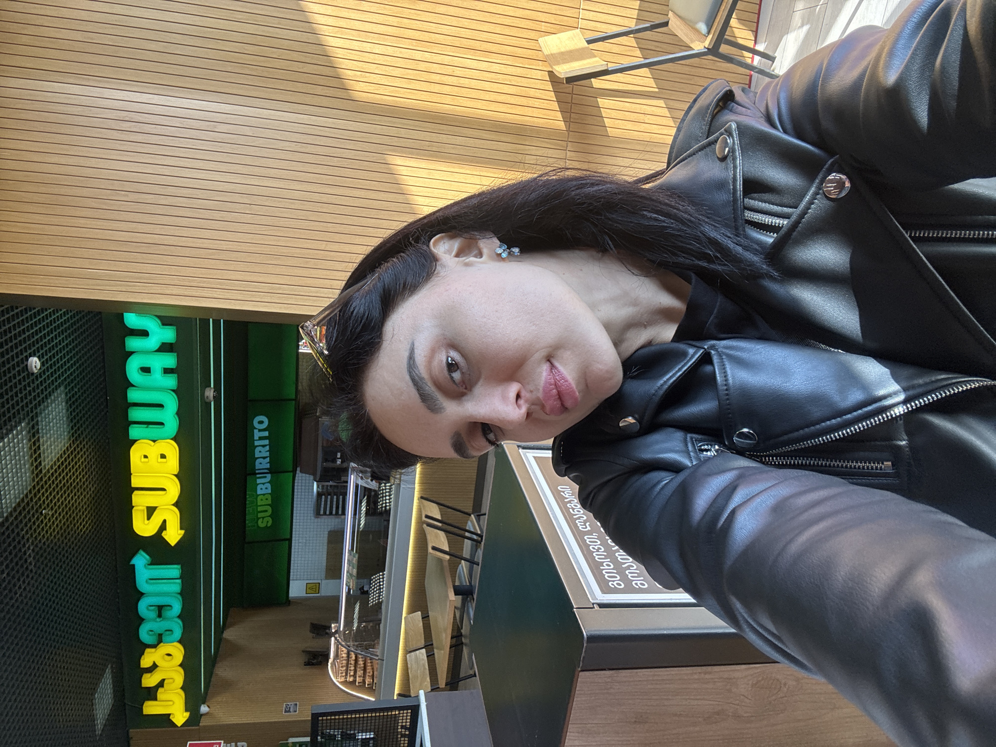 A person wearing a black leather jacket is taking a selfie inside a Subway restaurant in Tbilisi, Georgia. The Subway logo is visible in the background, along with tables and chairs. The person is facing the camera with a slight pout.