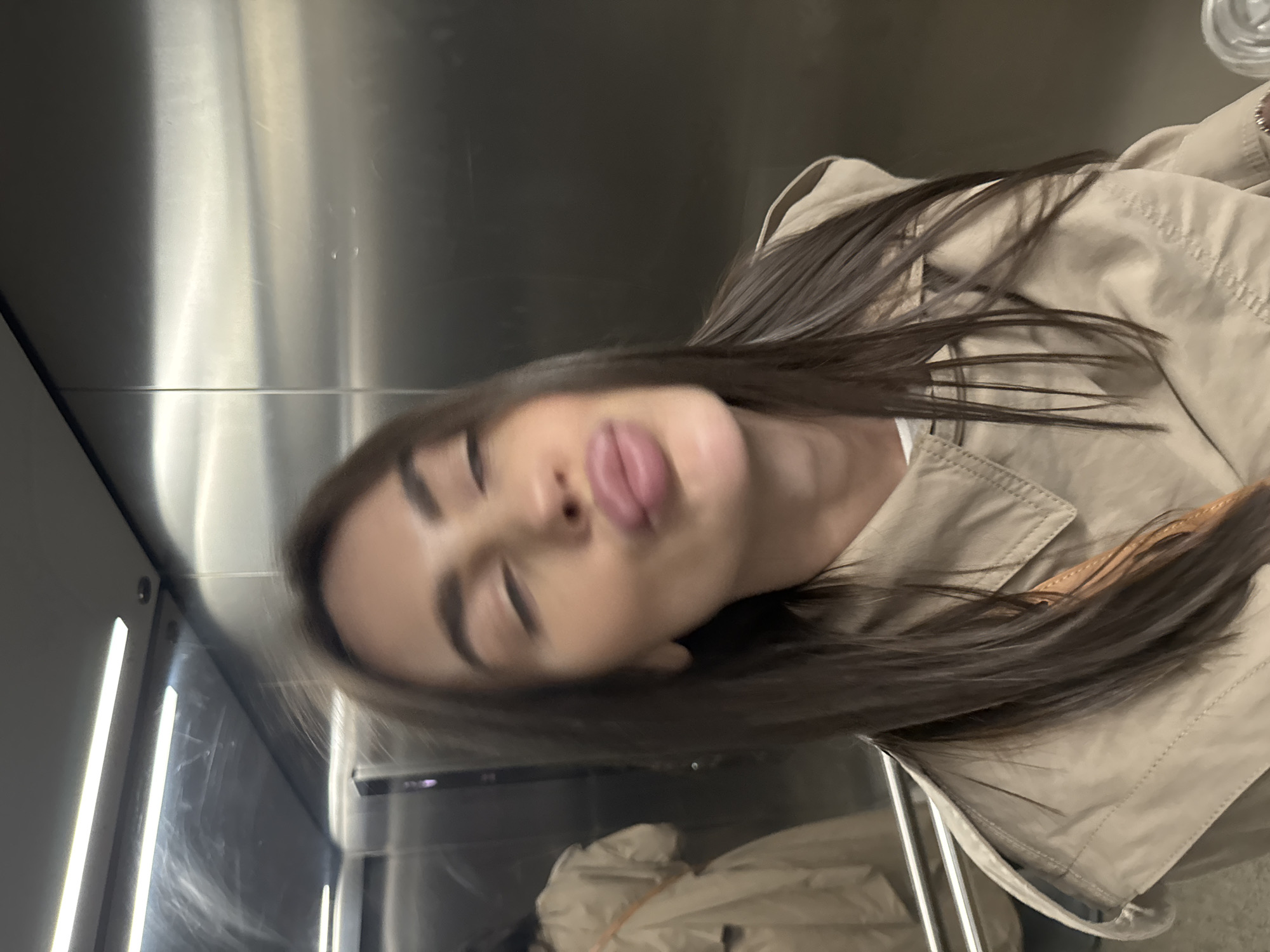 A blurry, rotated photo shows a woman with long, dark hair wearing a beige trench coat. Her eyes are closed, and her lips are pursed. The background appears to be the interior of a metal elevator. The location is unknown and cannot be determined from the image.