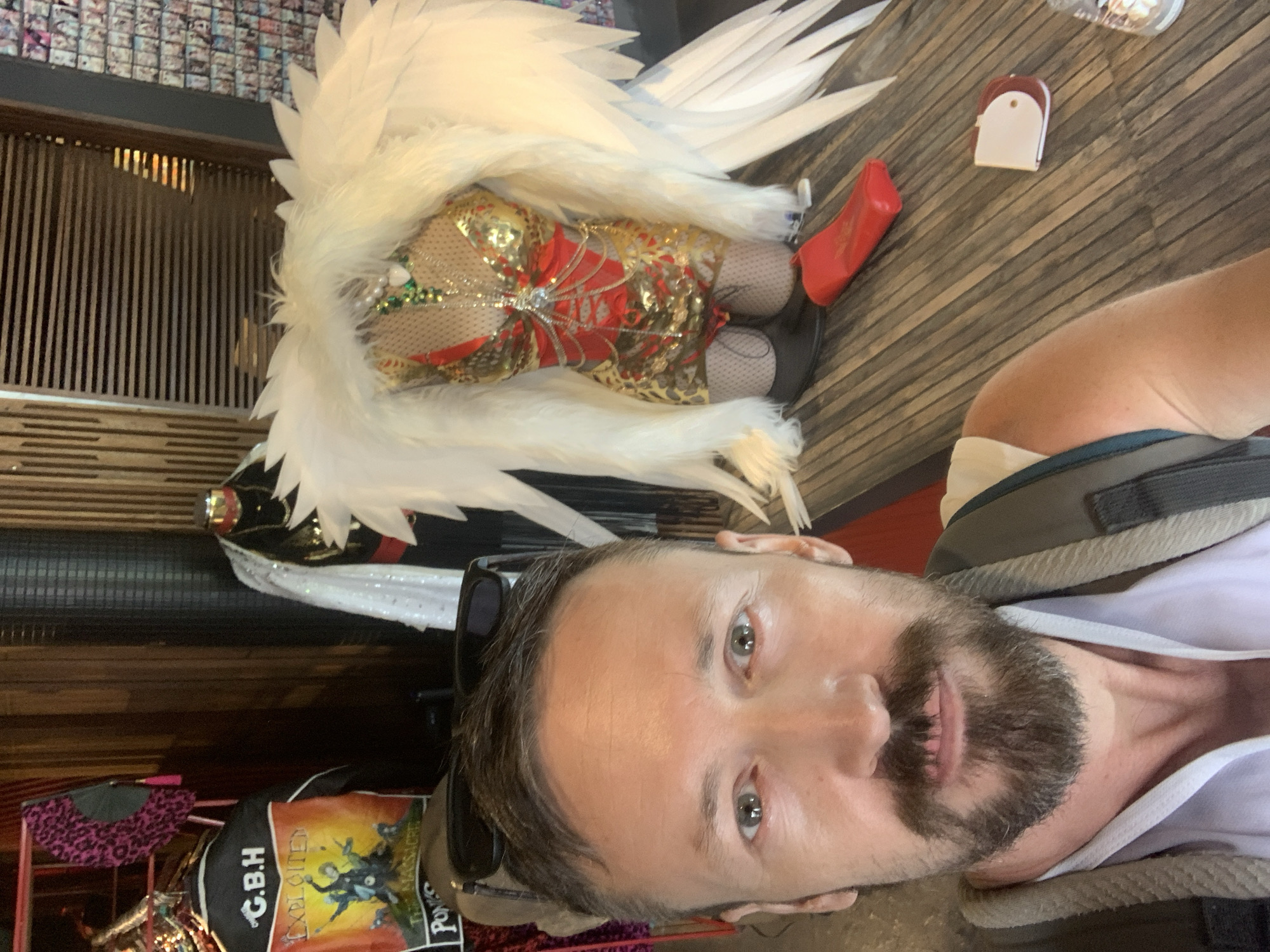 In the city of Ko Samui, Thailand, a man with a beard and sunglasses perched on his head takes a selfie in front of a display. The display features a mannequin adorned in a shimmering gold and red outfit, complete with large white feathery wings. To the left of the mannequin, a black jacket with the text "G.B.H," "Exploited," and "The Massacre" is visible. The background shows wooden paneling and what appears to be a wall covered in small items, possibly for sale. A red clutch bag rests on the wooden table in front of the mannequin. The man is wearing a light-colored shirt and a backpack.