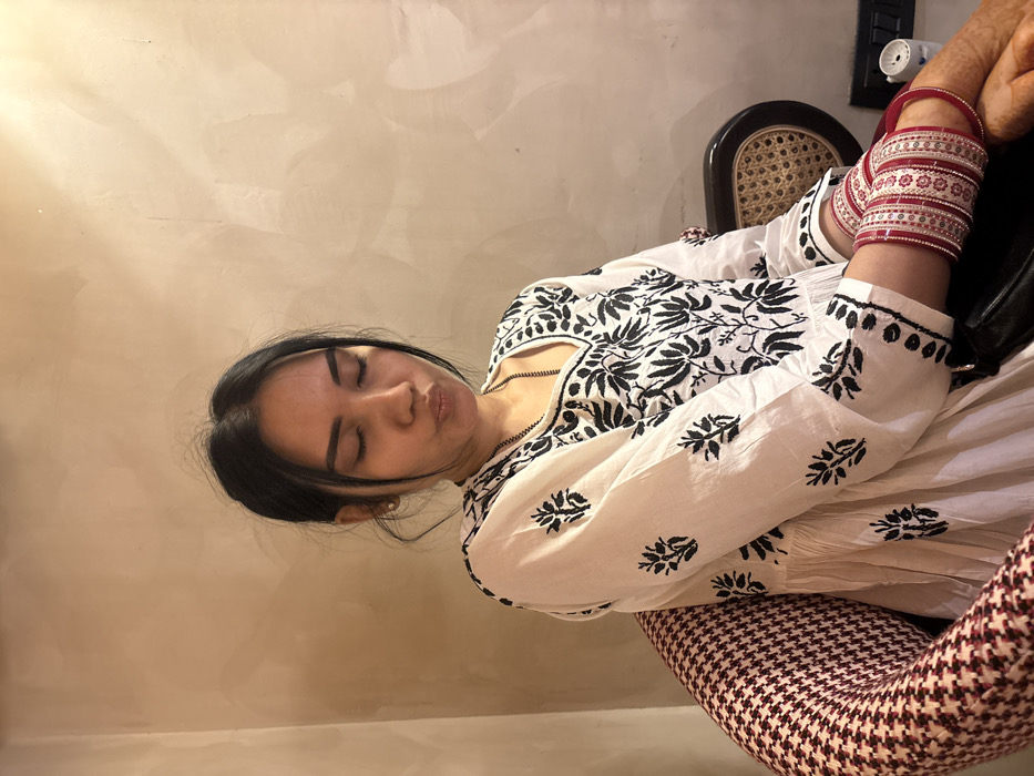 The image displays a female individual seated indoors in Howrah, India. She is shown from the chest up, with her dark hair pulled back, eyes closed, and lips pursed. A small stud earring is visible in her left earlobe. She wears a white, full-sleeved tunic featuring extensive black floral embroidery. A thin, dark chain is visible around her neck. Her left forearm is adorned with multiple red and white bangles, and henna is present on her hands. She occupies a chair with a red and white geometric patterned armrest visible on the left. A dark bag rests on her lap. To her right, a portion of a chair with a dark wooden frame and a cane back is visible. The background is a plain, light beige textured wall. In the lower right, a dark surface holds a small white object and a black electrical outlet.