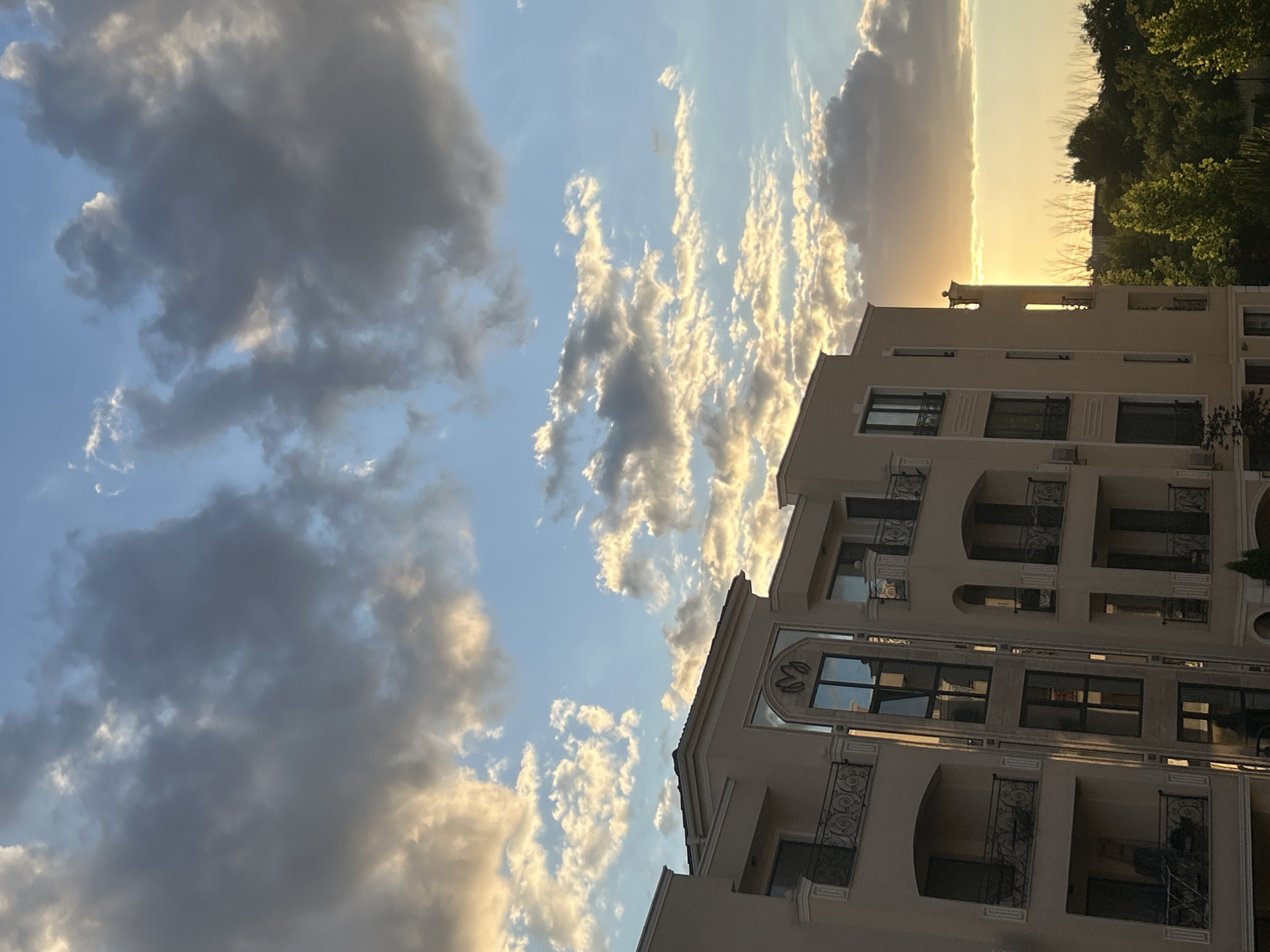 The image shows a modern apartment building in Plovdiv, Bulgaria, with balconies and large windows. The building is a light beige color and has a decorative facade with ornamental railings on the balconies. The sky above is filled with dramatic clouds, some dark and stormy, others lit by the warm glow of the setting sun. The sunset creates a golden horizon line that peeks through the clouds and illuminates the upper portions of the building. In the foreground, the tops of green trees are visible, adding a natural element to the urban scene. The overall impression is one of an approaching storm during a beautiful sunset over a residential area in the city.