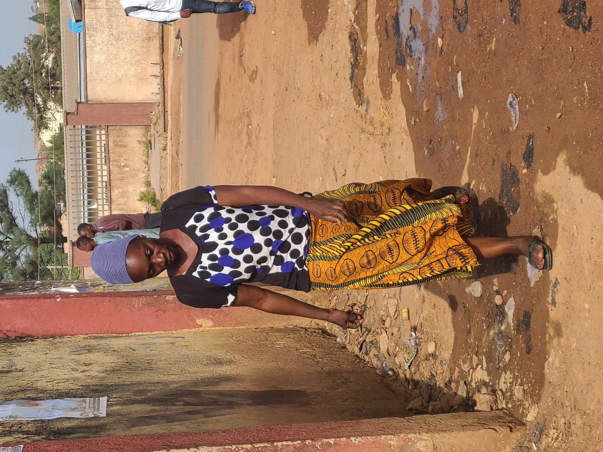 A woman lies on a street in Jos, Nigeria. She wears a head covering, a black and white polka dot shirt, and a yellow patterned skirt. The street appears to be dirty and muddy, with litter scattered about. Several other people are standing nearby. The environment includes buildings and trees in the background, as well as fences.