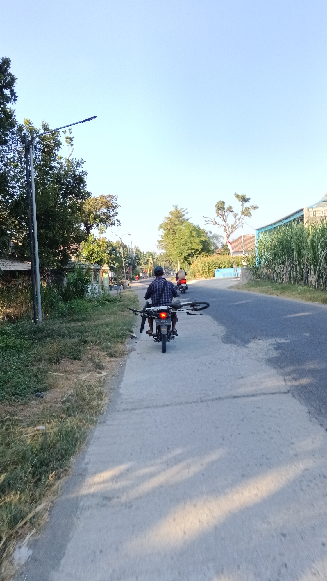 A person is riding a motorcycle on a paved road in Nambaan, Indonesia, carrying a bicycle on the back. The motorcycle has a license plate visible and appears to be black. The rider is wearing a plaid shirt and a dark cap. The bicycle is dark-colored, with its wheels extending outwards. Another motorcycle is visible further down the road, carrying a passenger. The environment is rural, with vegetation, trees, and buildings lining the road. The sky is clear and blue. A lamppost is visible on the left side of the road. The road surface is a mixture of concrete and asphalt. Sunlight casts shadows on the road.