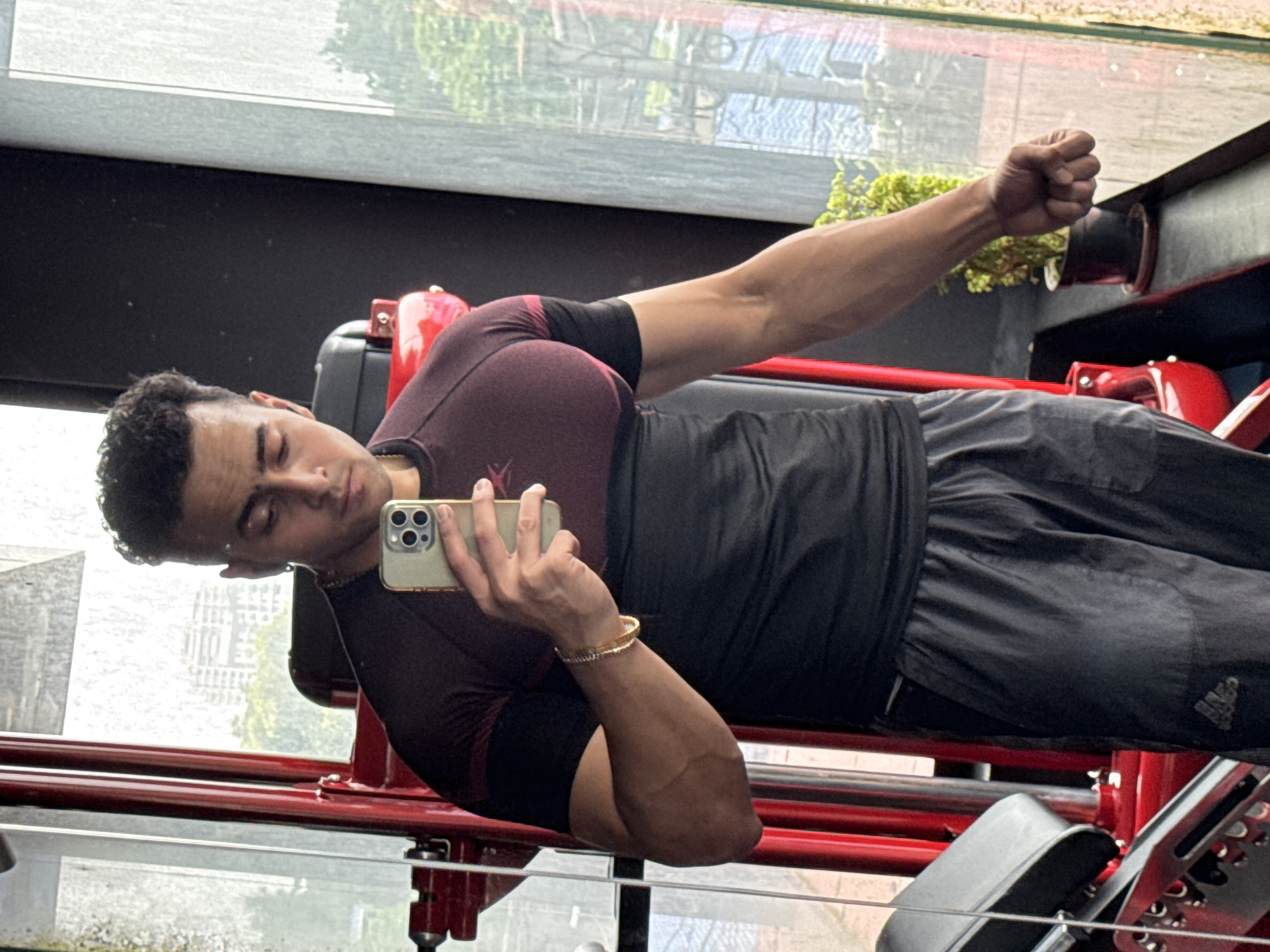 A young Indian man, appearing to be in his early twenties, is taking a mirror selfie in a gym. He has short, curly dark hair and is wearing a maroon and black athletic shirt and grey athletic shorts. A gold bracelet is visible on his left wrist. He is holding a smartphone in his right hand to take the photo. In the background, a red gym machine is visible, and through a large window, a cityscape with buildings and greenery can be seen, suggestive of Kolkata, India. The lighting indicates it is daytime.