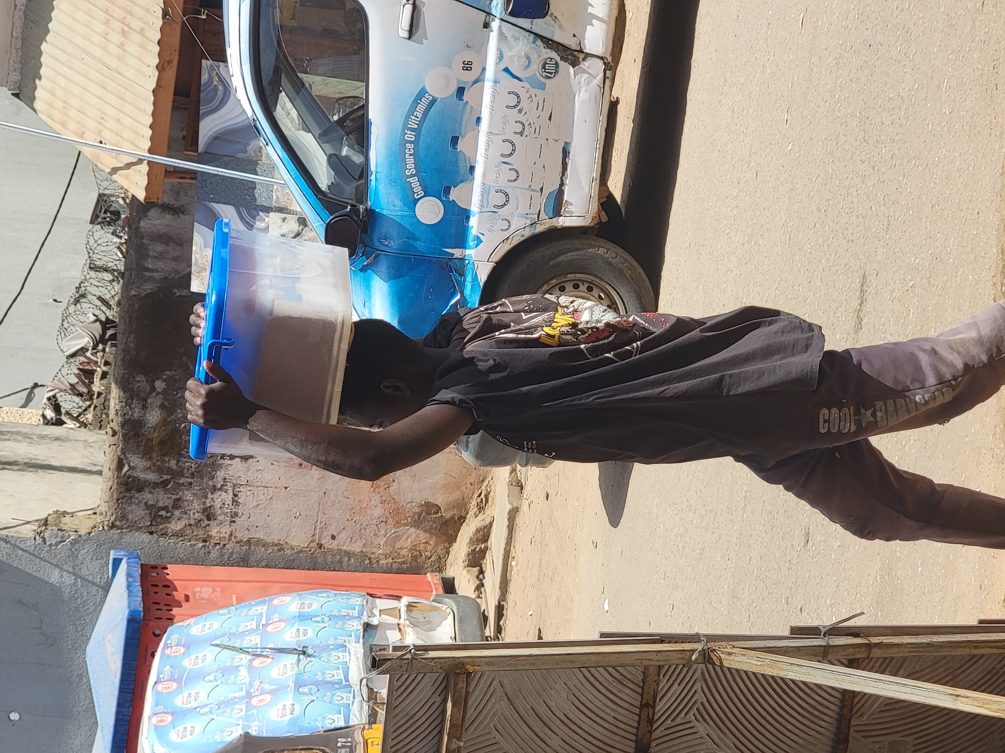 The image, taken in Maiburiji, Nigeria, depicts an outdoor scene under bright daylight. A person, likely male, with dark skin and dressed in a black t-shirt and dark pants, is actively transporting a large, clear plastic container with a blue lid, holding it over their head or shoulder. The container appears to hold a light-colored beverage.
To the right, a small commercial vehicle, painted blue and white, is parked. Its side features prominent branding with text including "Good Source Of Vitamins," "freshyo," "B6," and "Zinc," accompanied by graphics of beverage bottles. This vehicle is likely used for the distribution of these products.
In the lower-left, another blue container is visible next to a red cart or bin. This cart is filled with numerous stacked packs of what appears to be "Freshyo" brand beverages, with legible text like "Straw" and "Hole" on the packaging. The person's pants also show text, possibly "COOL★BARN." The background reveals concrete walls, some showing signs of wear, and a section of corrugated metal roofing. Barbed wire can be seen on a wall in the distance. The ground is a light-colored, paved surface, and distinct shadows indicate a sunny time of day. The scene suggests commercial activity, likely beverage sales or delivery, in a vibrant street or market setting.