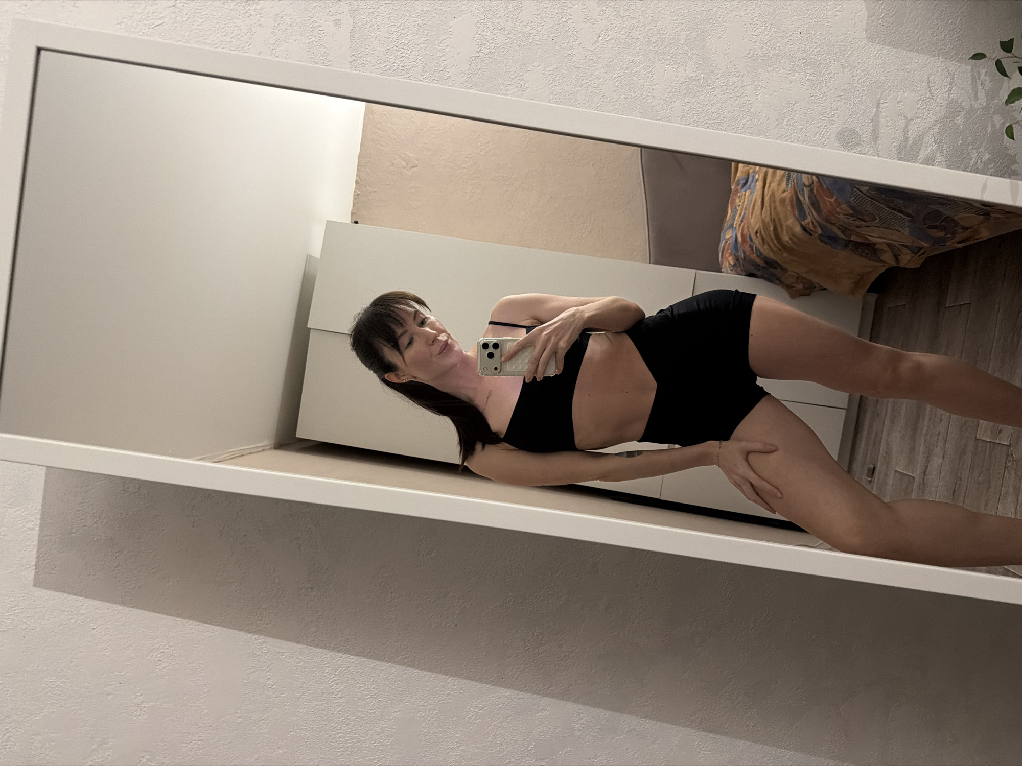 A full-body mirror selfie captures a woman indoors, rotated sideways. The woman, with dark hair and bangs, is wearing a black sports bra and matching shorts. She holds a light-colored smartphone (an iPhone with three visible camera lenses and a clear case) in front of her to take the photo, looking towards the phone's screen. Her left hand rests on her right thigh, and her body is slightly angled.
The setting, reflected in the large white-framed mirror, shows a room with light-colored, textured walls. Visible furniture includes a white dresser or cabinet behind the woman. In the upper right corner of the mirror's reflection, a patterned pillow or blanket on what appears to be a bed can be seen, along with a glimpse of a green houseplant. The floor visible in the reflection has a wood or laminate texture. The lighting suggests an indoor, possibly evening or night-time setting, given the shadows cast. No discernible text is visible in the image. The photo was taken in Perm, Russia.