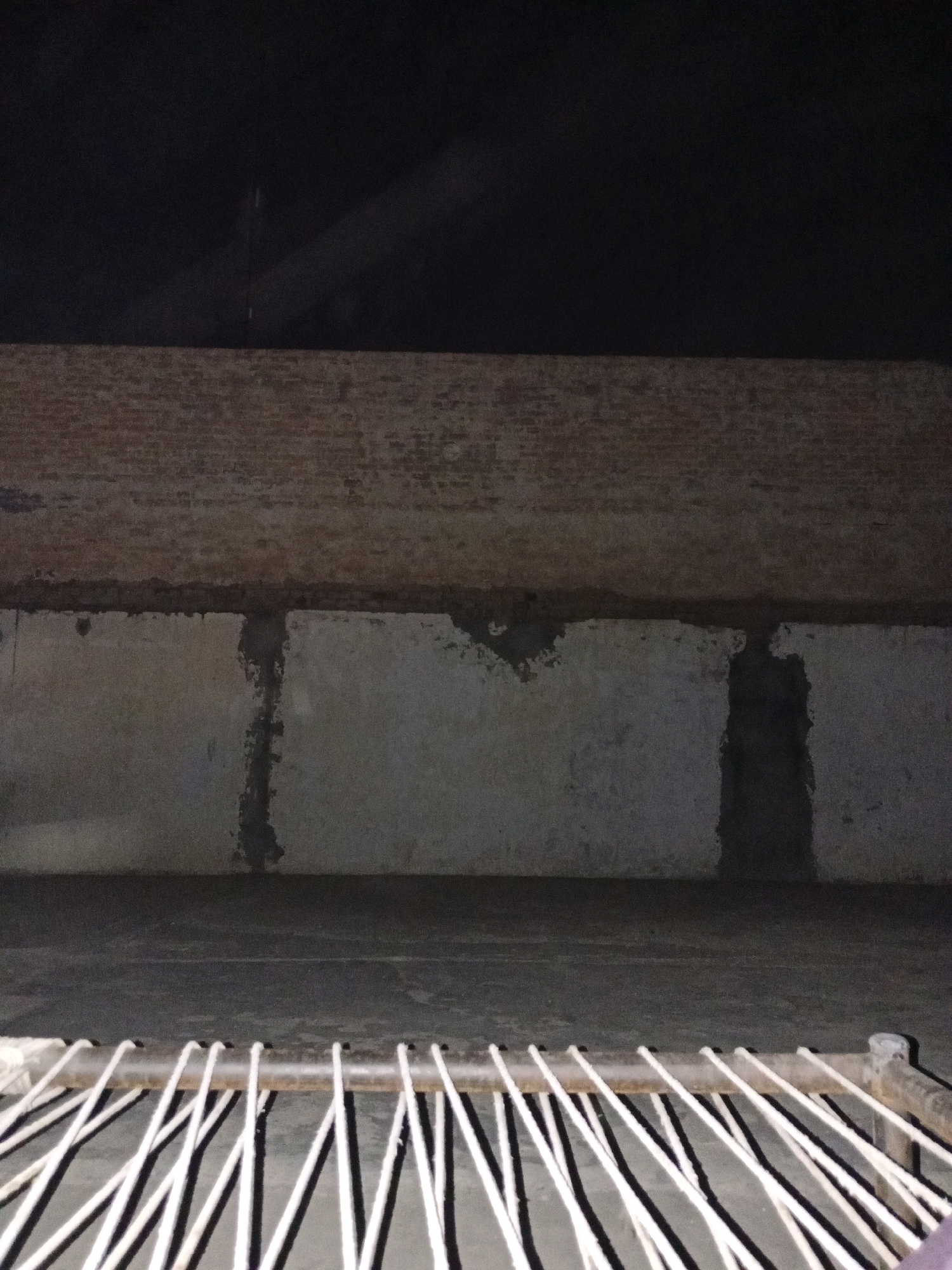The image depicts an indoor scene, likely within a structure in Bahawalpur City Tehsil, Pakistan. The foreground shows a close-up view of a metal bar supporting multiple white cords that are spaced evenly apart. These cords extend downwards and to the right, creating a striped pattern with shadows cast on a dark grey floor or surface.
Behind the cords, the middle ground reveals a wall constructed of grey concrete blocks, with visible mortar lines and patches of rough texture. A horizontal dark band runs across the top of this concrete section, suggesting a separation between wall layers or a shelf. Above this band, the background shows a wall made of aged bricks, exhibiting signs of wear and a rough, textured surface. The uppermost section of the image is predominantly dark, with a faint diagonal light source visible in the upper right quadrant, casting a subtle illumination on the dark surface.
The overall impression is that of a simple, utilitarian space, possibly a storage area, workshop, or even a constructed enclosure like a shed or a room within a larger building. There are no people or animals visible in the image, and no overt actions or interactions are taking place. The environment appears to be dimly lit, with the primary light source suggested by the diagonal beam in the background. The state of the walls and the presence of the corded structure suggest a functional rather than decorative purpose for the space.