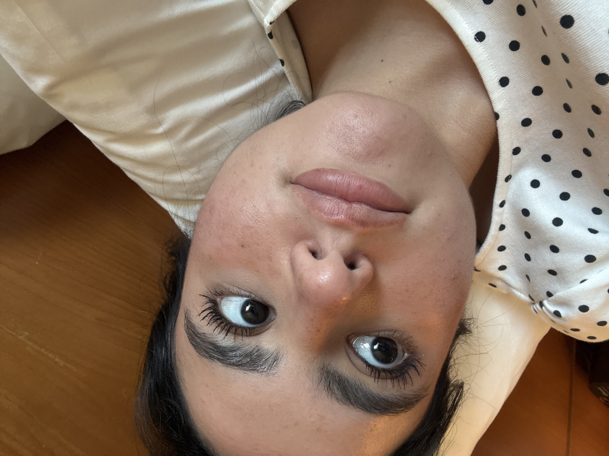 A close-up, high-angle selfie shows a young woman lying in bed with her head propped on a pillow. Her eyes are wide and looking in different directions, giving her a surprised or playful expression. She has dark hair and full eyebrows, and her eyelashes are noticeable. Her lips are slightly parted. She is wearing a white top with black polka dots. The background consists of a beige pillow and a wooden headboard. The image appears to have been taken indoors, possibly in a bedroom. The location is not specified.