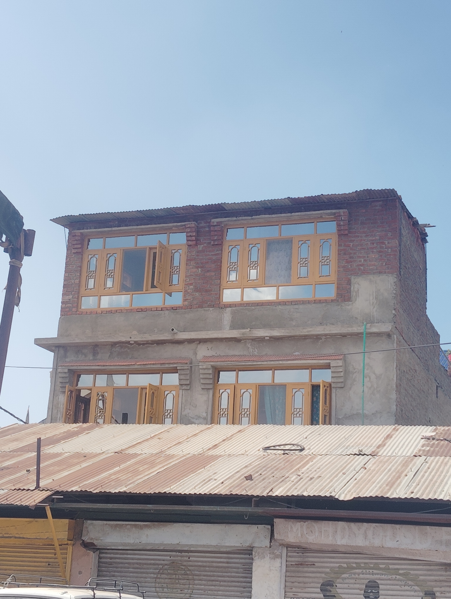 This is a medium shot, outdoor, eye-level, vertical, daylight photograph of an unfinished building under a clear blue sky in Srinagar, India. The building has three visible levels. The top level is constructed of red brick, with two large, ornate wooden-framed windows. The middle level is made of concrete with two similar wooden-framed windows. The bottom level, partially obscured by a corrugated metal roof, also features wooden-framed windows and doors. The corrugated metal roof is rusty and appears to cover commercial storefronts below. A metal pole and scaffolding are visible on the left side of the image.