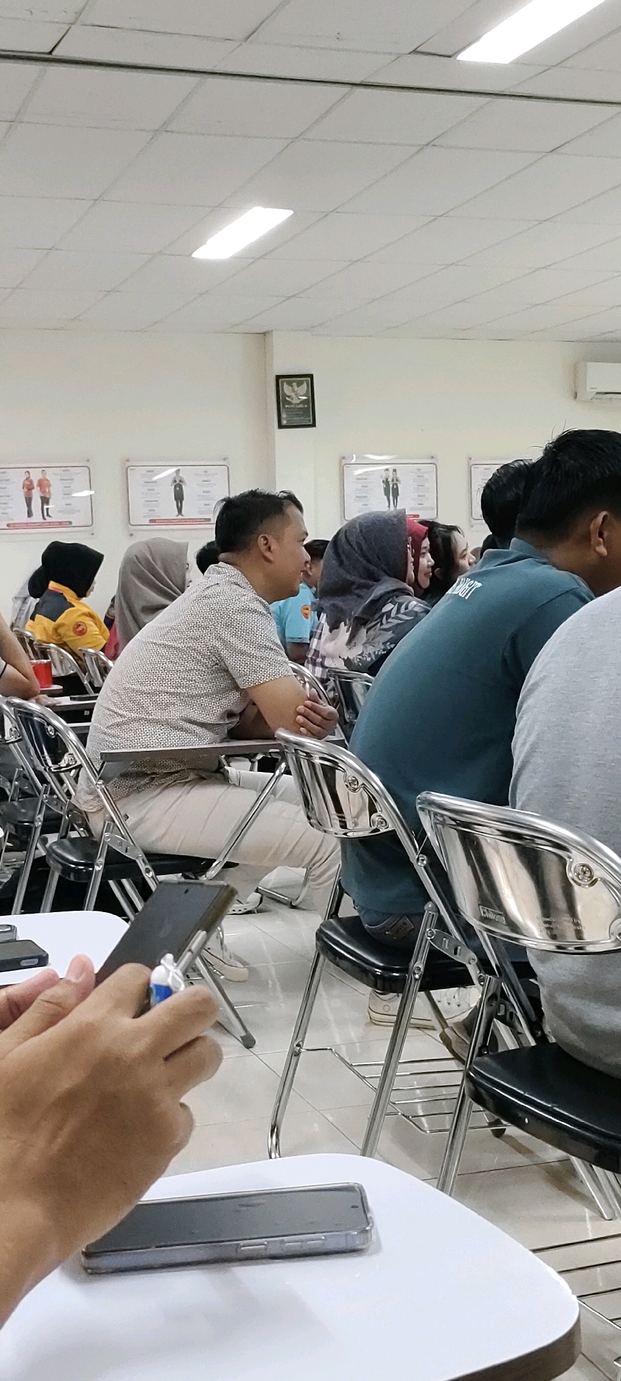 The image depicts an indoor scene, likely a classroom or lecture hall, with multiple people seated in chairs. The room has a white tiled ceiling with fluorescent lighting panels. Several framed posters with images and text are displayed on the back wall. In the foreground, a person's hands are visible, holding a smartphone, and another smartphone is placed on a white table. The seated individuals are a diverse group, with some wearing headscarves. The chairs are metal-framed folding chairs with black seats and backs. The overall atmosphere appears to be a gathering or educational event. There is no visible indication of the city of Nusa Indah, Indonesia, in the image itself.