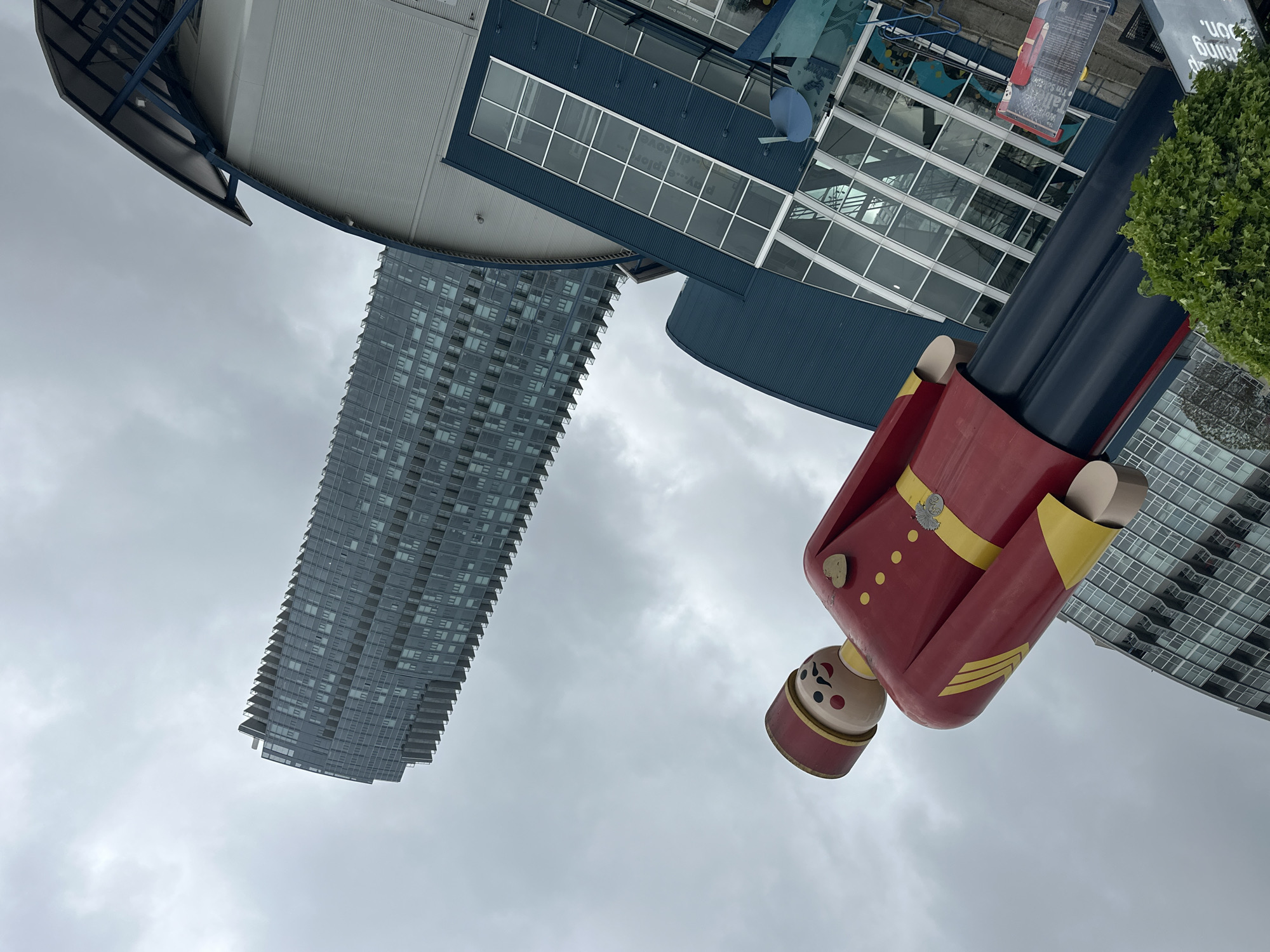 An outdoor urban scene is depicted, centered around a large, colorful statue of a toy soldier or nutcracker figure against a backdrop of modern buildings under a cloudy sky. The prominent statue, situated on the right side of the frame, is predominantly red and blue with yellow accents. It wears a red jacket detailed with yellow trim, gold buttons, and a yellow belt featuring a silver-colored emblem. Its trousers are blue, and it is topped with a red and yellow hat. The face is painted with black eyes, red cheeks, and a small mouth.
In the middle-left background, a tall, slender high-rise building, composed of many glass windows and balconies, ascends towards the sky. Another building with a distinct blue roof and large glass facades is visible behind and to the upper left of the statue. Text on this building's windows or signage includes partial phrases such as "play...explor", "the quay side", "Walk the", "I'm So", and "Fun. building on.". A patch of green foliage, likely a bush, is visible in the upper right corner. The sky is uniformly grey and overcast, indicating a sunless daytime. No human activity is present in the static scene.