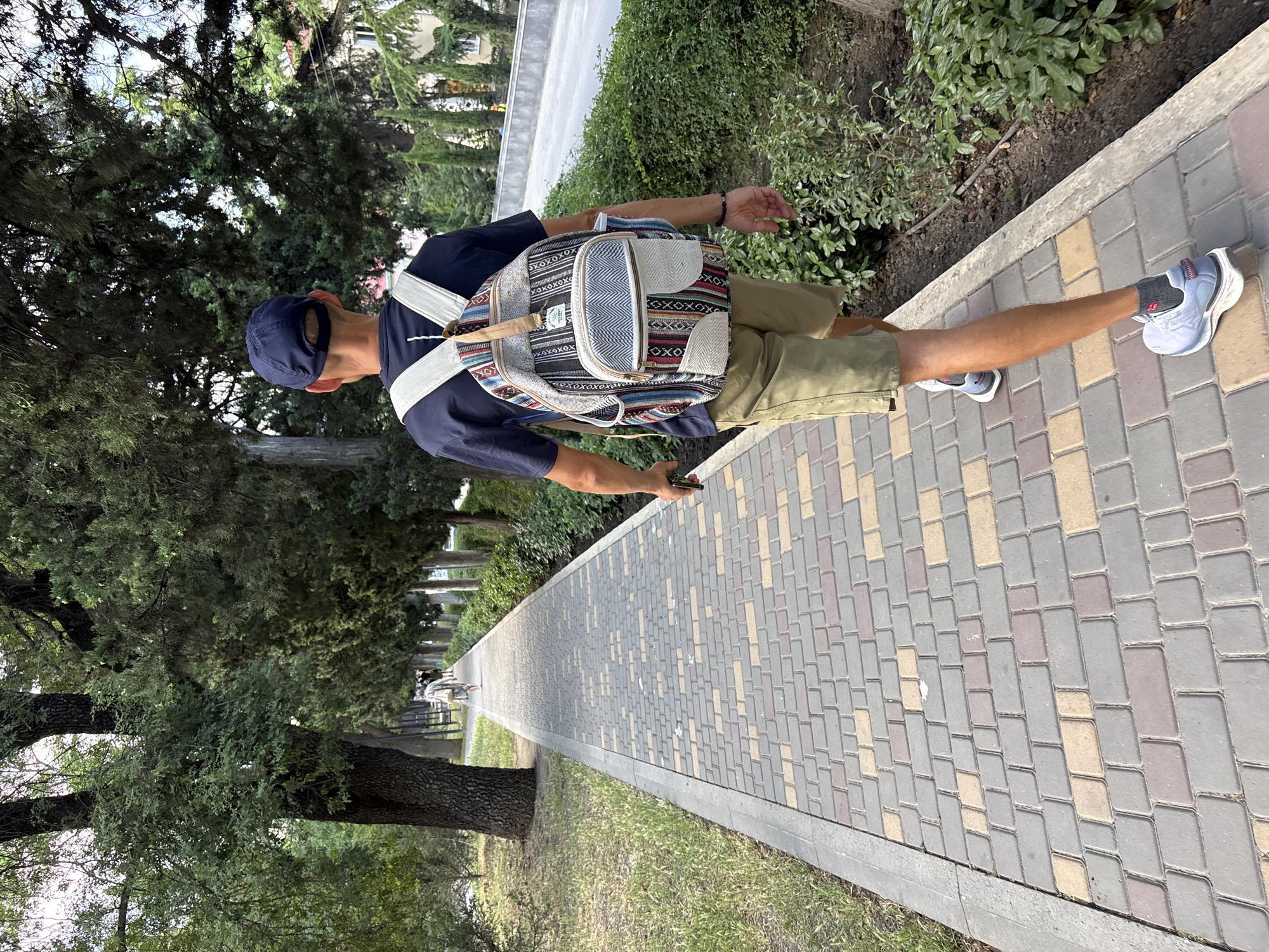 A young man with a backpack walks away from the camera on a paved walkway lined with trees and bushes. He is wearing a dark blue t-shirt, khaki shorts, a navy blue baseball cap, and athletic shoes. The walkway is constructed of grey, beige, and reddish-brown rectangular pavers arranged in a repeating pattern. To the left of the walkway, there is a grassy area with a large tree trunk. To the right, there are green bushes and a low concrete curb. In the background, a paved road with cars can be seen. The scene appears to be a park or a promenade. The man is holding a mobile phone in his right hand. The location is identified as being in the city of Alushta, Ukraine.