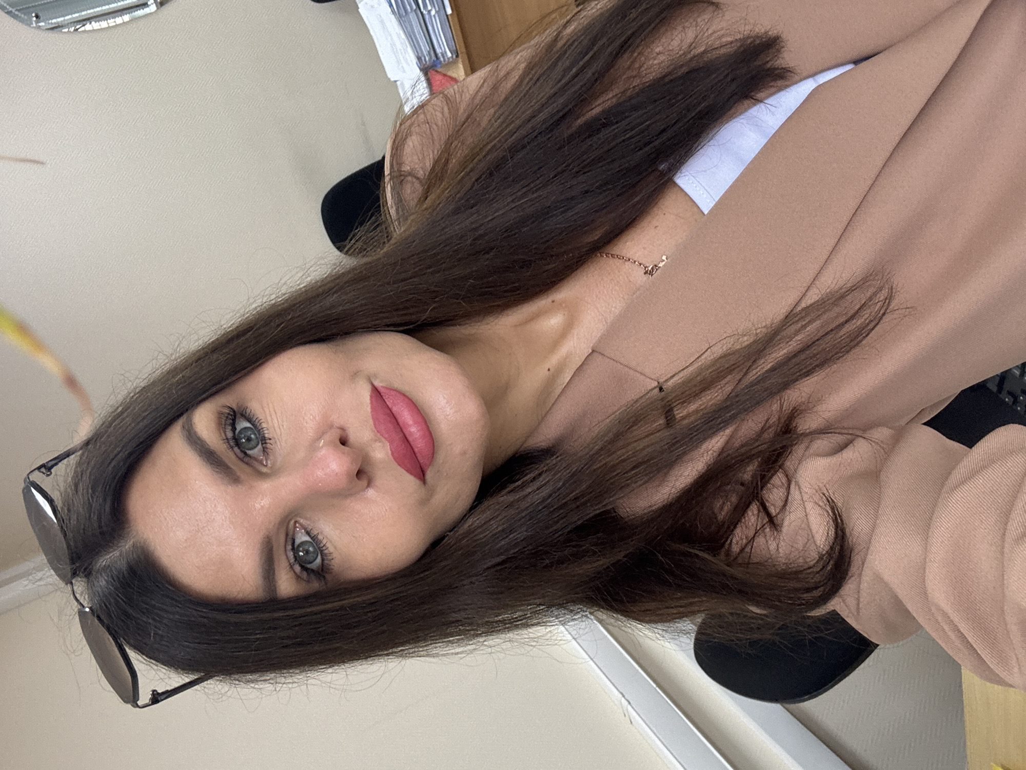 A young woman with long, dark brown hair is captured in a close-up, slightly over-the-shoulder selfie. She is wearing a light brown blazer over a white top and a delicate rose gold necklace. Her makeup is noticeable, with defined eyebrows, mascara, and a bold pink lipstick. She is also wearing dark-rimmed sunglasses perched on her head.
The background is blurred, but hints of an office environment are visible. A desk with some stationery items, including what appears to be a stapler, is partially visible to the left. To the right, a part of a chair can be seen. The lighting suggests an indoor setting, likely during the daytime. The woman's expression is neutral, with her gaze directed towards the camera. The image is taken with the camera rotated, giving it a sideways orientation. The overall scene suggests a casual moment captured in a professional or study setting, possibly in Chaikovsky, Russia.