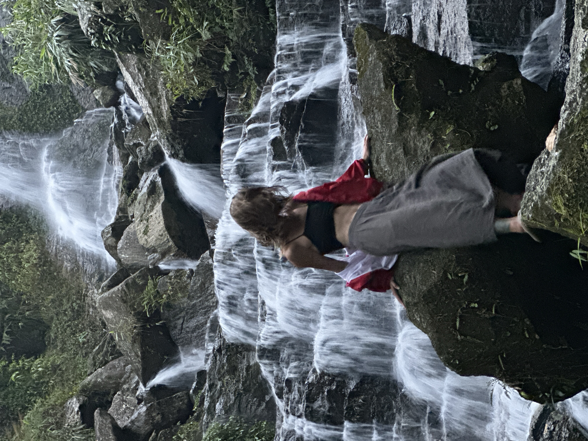A woman is depicted lying horizontally on a large, moss-covered rock formation next to a cascading waterfall in Thông Nguyên Commune, Vietnam. The image, if rotated to its natural orientation, shows the waterfall descending vertically. The woman has long, light brown hair and is wearing a dark-colored top and grey pants, with a vibrant red garment (possibly a shawl or light jacket) draped around her midsection. Her pose suggests she is either resting or engaging in a photoshoot, with her body stretched along the rock and her head positioned close to the flowing water.
The background is dominated by a powerful, multi-tiered waterfall, where water streams over dark, layered rock formations, creating a frothy, white descent. Lush green foliage, including various plants and ferns, grows abundantly on the rocks and along the periphery of the waterfall, indicating a dense, natural environment.
The lighting appears soft and diffused, characteristic of daytime under an overcast sky or within a shaded canyon, as there are no harsh shadows. The atmosphere is one of serene natural beauty and tranquility. There is no visible text in the image. The scene captures an individual engaging with and enjoying the breathtaking natural landscape.