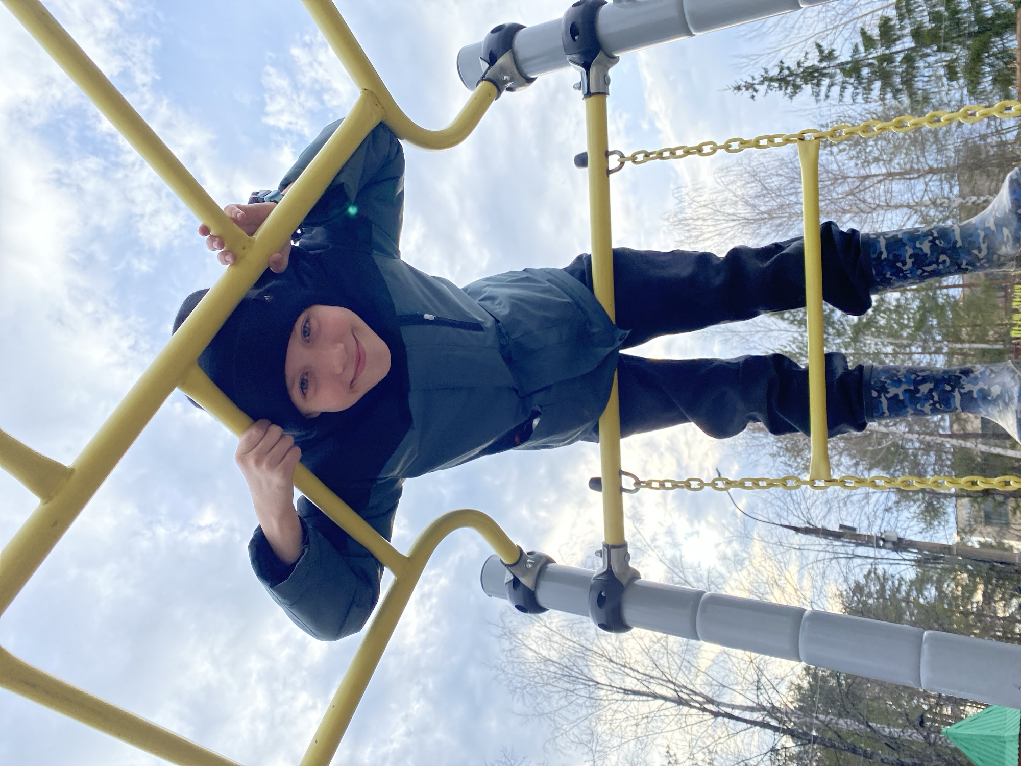 A young child is captured in a playful moment, hanging upside down from bright yellow playground equipment against an overcast sky. The child, wearing a dark winter jacket, black pants, a black beanie, and distinctive blue-grey camouflage rubber boots, grins broadly at the camera. Their hands firmly grip the yellow metal bars, which appear to be part of a monkey bar or ladder structure.
The image is taken from a low angle, looking directly upwards, enhancing the sense of the child defying gravity. The sky above is a light grey-blue with scattered white clouds, indicating an overcast but well-lit daytime. In the background, beyond the playground structures, a landscape of bare deciduous trees and a few evergreen trees suggests a colder season, such as late autumn, winter, or early spring. Other parts of the playground equipment, including a grey horizontal bar with black connectors and a segment of a yellow plastic chain, are also visible. The child's happy expression conveys a sense of joy and adventure during outdoor play. No visible text is present. The scene is set in Chaikovsky, Russia.