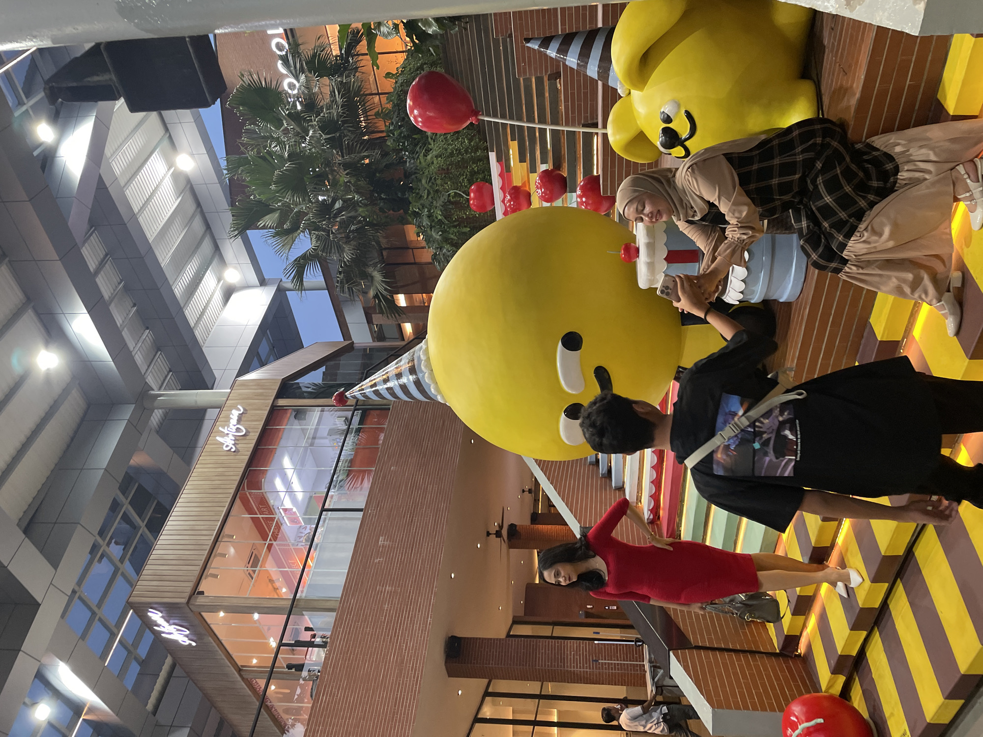 The image depicts a group of people posing with large, cartoonish yellow characters wearing birthday hats and standing near a cake. One person is holding a smartphone and taking a photo of another person with the character. The environment is an outdoor space that appears to be a pedestrian area adjacent to a building with glass windows and brick sections. The building displays the name "Soyoum" in a stylized font. Palm trees and decorations like red balloons are visible, suggesting a festive occasion. The scene suggests a promotional event or a whimsical public art installation. Given that Soyoum is a restaurant located in North Jakarta, Indonesia, it is likely that the image was taken in the area. The person in the red dress stands a bit away from the main interaction.