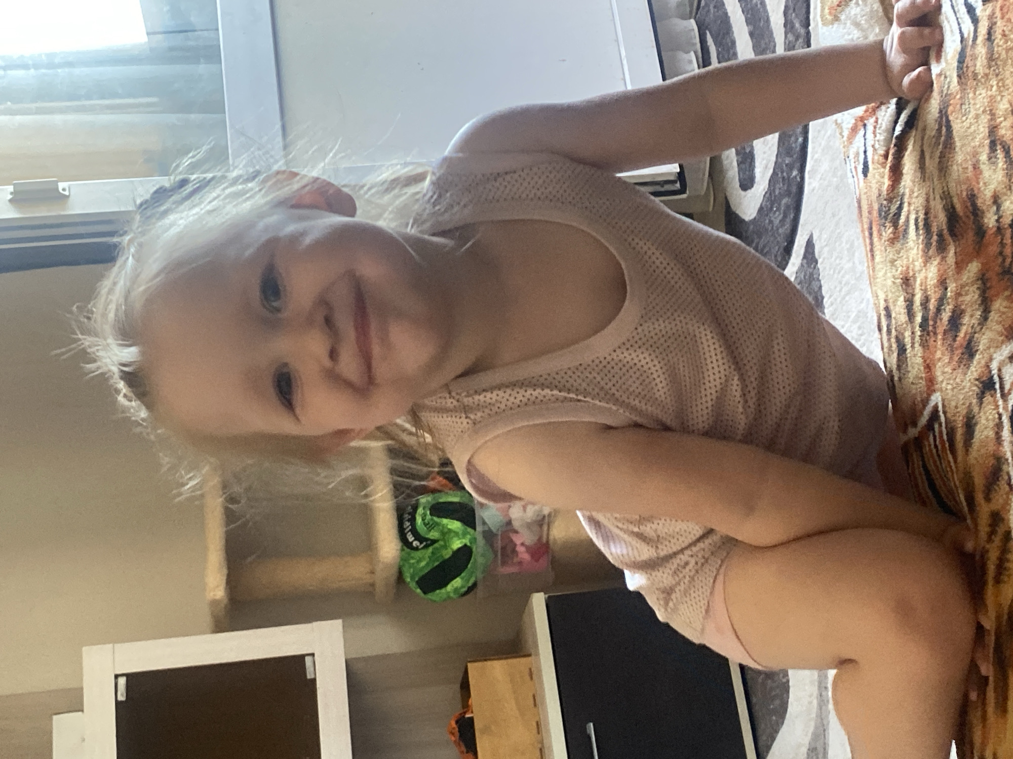 A young girl with blonde hair is leaning towards the camera, her head tilted. She is wearing a light pink sleeveless top and pink bottoms. Her left arm is extended, and she is holding onto a patterned blanket with a tiger-like print. In the background, a green ball with black markings is visible, along with some other objects that appear to be toys or craft supplies. The setting seems to be indoors, possibly a living room or playroom, with furniture and shelves visible. The lighting suggests it could be daytime. The girl's expression is playful and engaging.