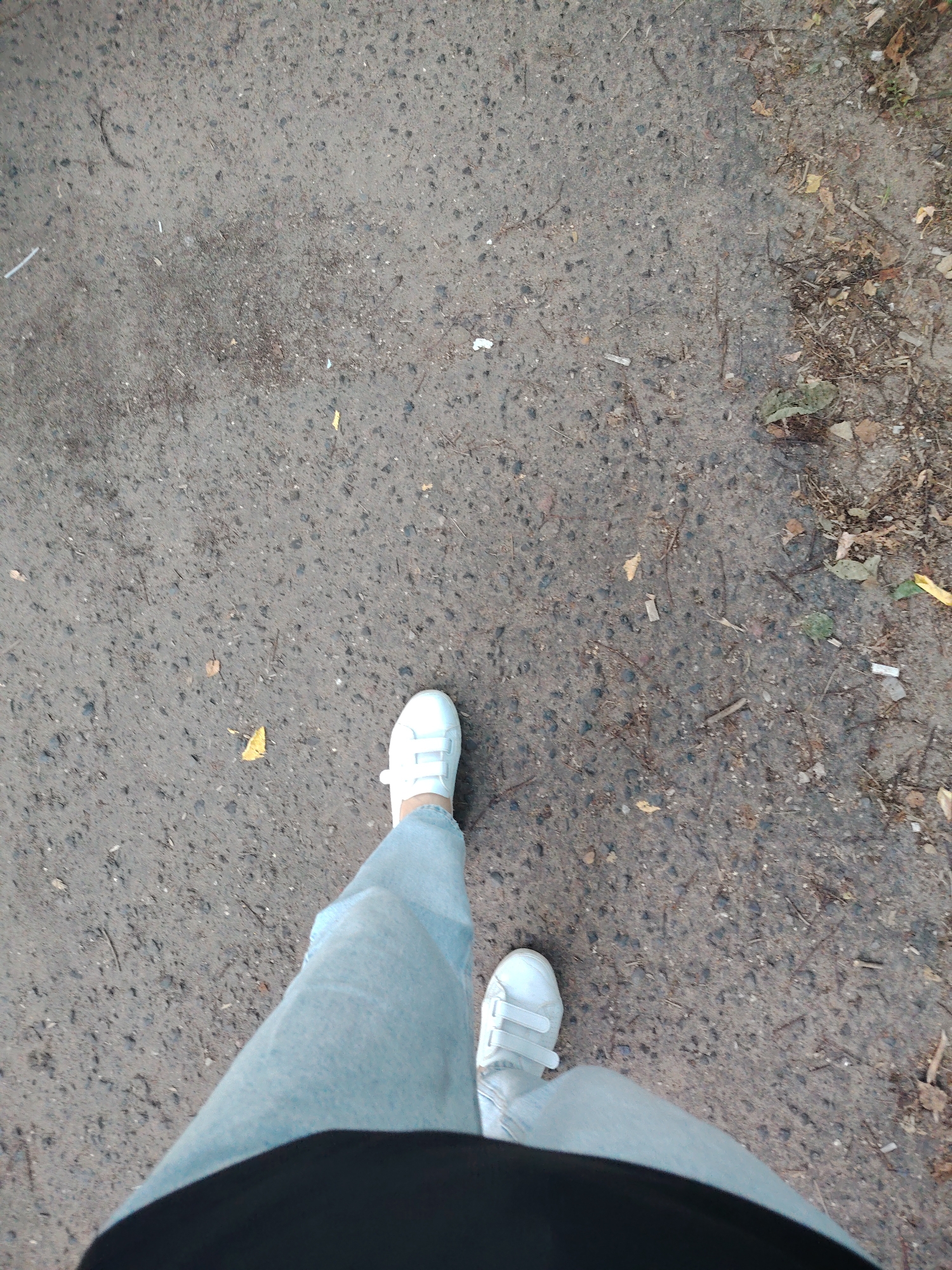 This is a first-person perspective shot of a person wearing light blue jeans and white velcro sneakers walking on a paved surface in Konakovo, Russia. The ground is gray asphalt with scattered small stones, debris, and fallen leaves, suggesting an outdoor setting possibly in a park or on a street. The lighting is overcast, casting minimal shadows. The focus is on the lower legs and feet of the individual.