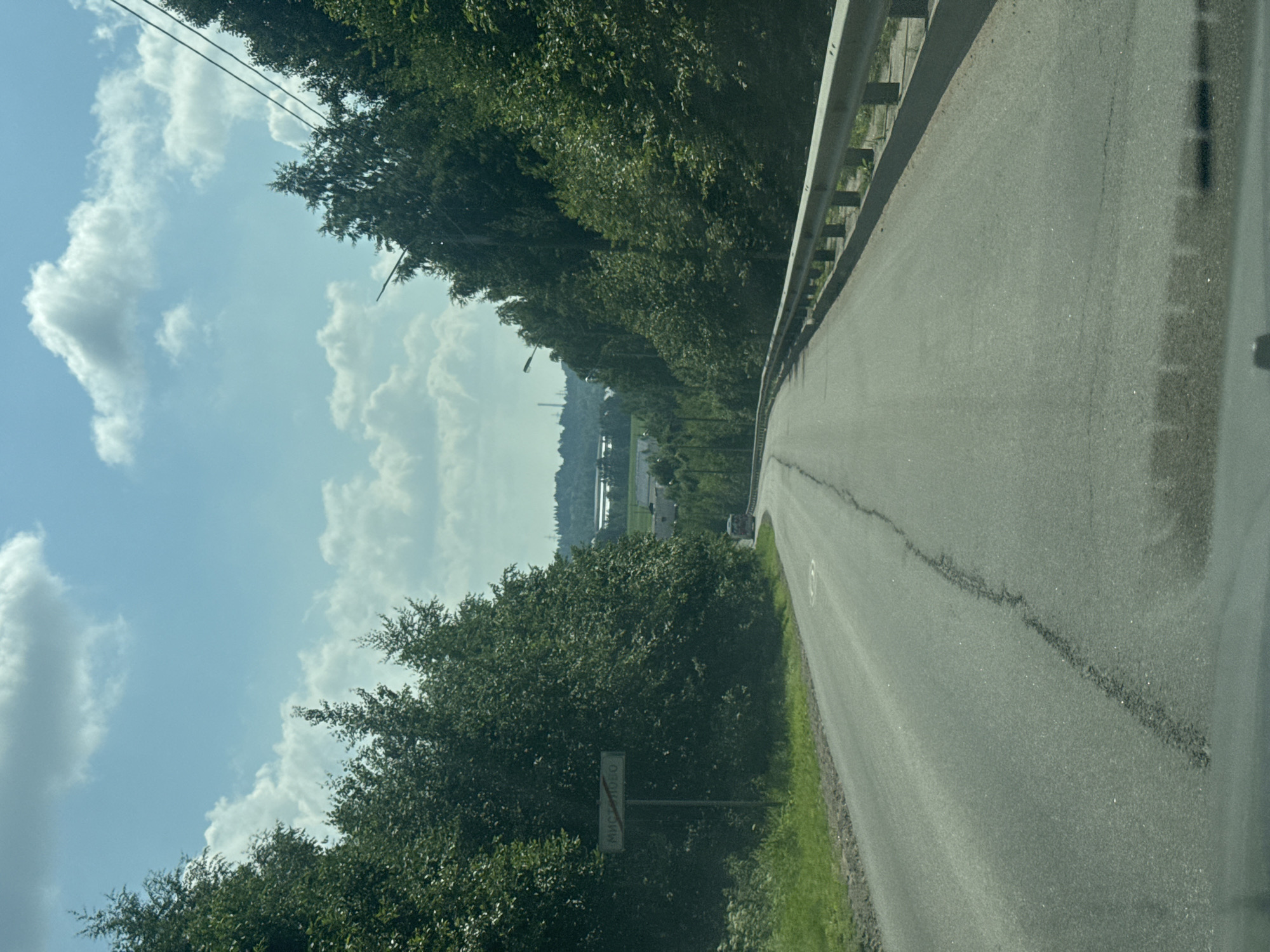 This media file captures a view from a vehicle traveling on a paved road. The road curves to the right, bordered by a metal guardrail on the left and a grassy shoulder. Dense green trees line the left side of the road, creating a canopy that extends into the background. In the distance, beyond the trees, a hazy landscape is visible with hints of industrial structures and a body of water. The sky is a clear blue with scattered white cumulus clouds, suggesting a sunny day. A street lamp is visible among the trees on the left side of the road. A road sign with Cyrillic text, "ГОРОДОК", is partially obscured by foliage on the left. The overall impression is one of a scenic drive through a rural or semi-rural area. The time of day appears to be daytime, given the bright sunlight and clear sky. The weather is fair and pleasant. The activity is the act of driving, as implied by the perspective from inside a vehicle.