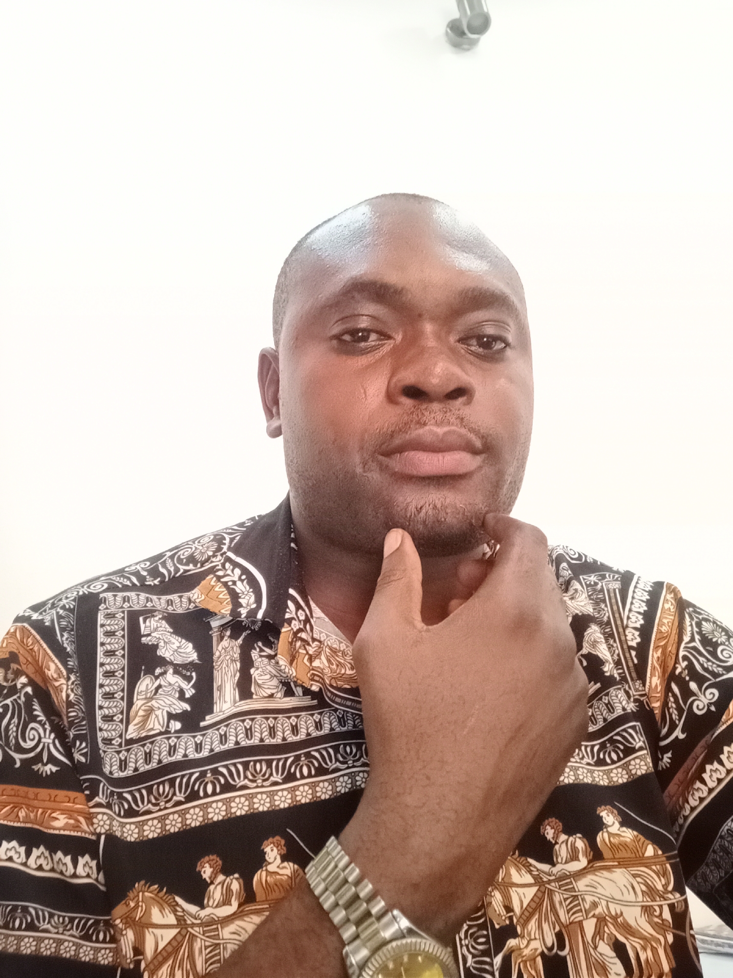 A man, appearing to be in his 30s or 40s, is captured in a close-up portrait, likely a selfie, in an indoor setting. He has a shaved head and a short beard or stubble on his chin, and looks directly into the camera with a neutral, engaging expression. His right hand is positioned with fingers resting on his chin, suggesting a thoughtful or pensive pose.
He wears a short-sleeved button-up shirt featuring an intricate pattern of black, gold, and white. The design includes classical motifs such as figures in chariots, people in traditional attire, and decorative borders. On his left wrist, he has a silver-toned watch with a metallic link band and a gold-colored watch face.
The background is a plain, light-colored wall, providing a simple and uncluttered backdrop. In the upper right corner, a small, metallic fixture or pipe is partially visible. The lighting is bright and even, consistent with an indoor environment. The scene is likely set in Gwarinpa, Nigeria. There is no visible text in the image.