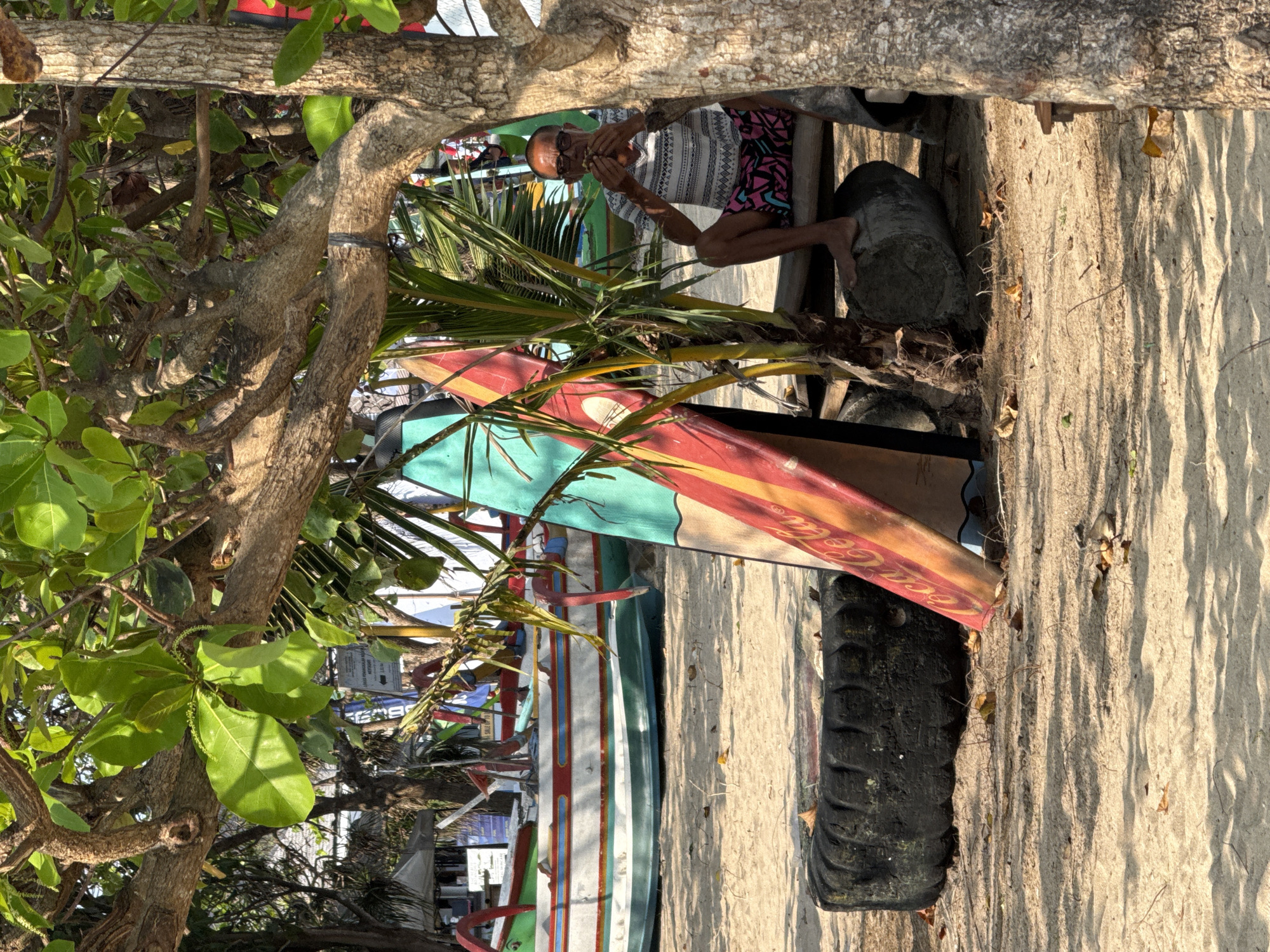 The image depicts a sunny beach scene in Denpasar, Indonesia, likely on Sanur Beach due to the presence of traditional jukung boats. In the foreground, two colorful surfboards lean against a tree. To the left, a large, worn tire serves as a makeshift seat or barrier. A man, wearing sunglasses, a patterned t-shirt, and patterned shorts, sits on a wooden bench made of logs, holding something to his face as if eating. Behind him, several traditional Balinese fishing boats with outriggers are pulled up on the sand, suggesting a typical fishing village setting. The environment is sandy with scattered leaves and dappled sunlight filtering through the trees, indicating a warm, tropical atmosphere.
