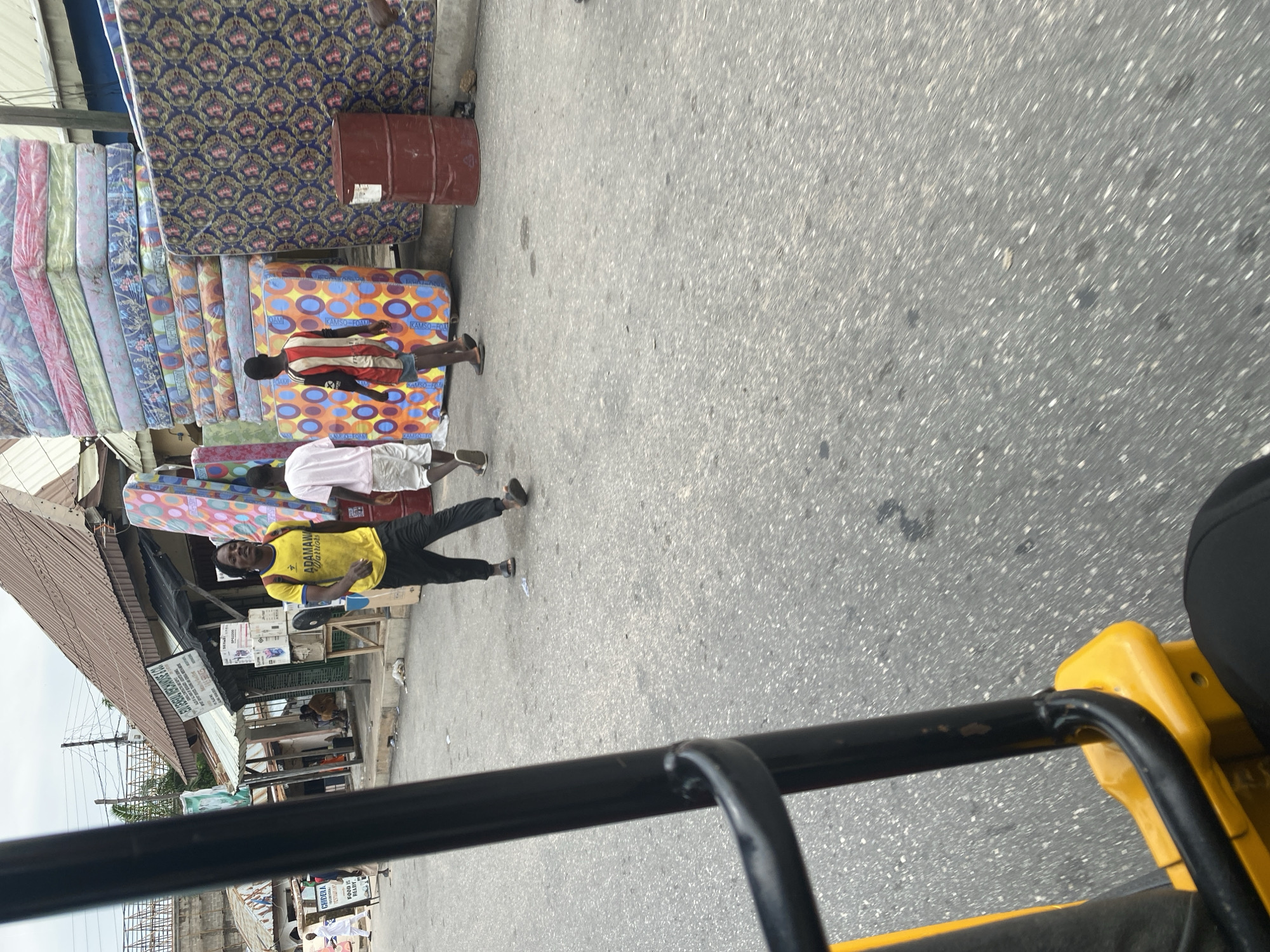 The image shows a street scene in Jalingo, Nigeria, with a focus on a mattress shop. Several mattresses are stacked vertically, some wrapped in plastic and others with colorful patterned covers. A few people are visible in the scene. In the foreground, a person wearing a yellow t-shirt with "ADAMAWA WARRIORS" printed on it and black pants is standing and gesturing. Another person in a pink t-shirt and white shorts is also present, along with a third person whose body is partially visible. A large, reddish-brown barrel stands near the mattresses. The ground is paved asphalt. In the background, there are various market stalls and buildings with corrugated metal roofs. The overall impression is that of a busy street market. The time of day appears to be daytime, and the weather seems overcast. The image is captured from a low angle, possibly from a moving vehicle, as suggested by the blurry foreground element which appears to be part of a vehicle. Visible text includes "ADAMAWA WARRIORS" on the yellow t-shirt and signs on the market stalls, though the text on the signs is largely illegible.