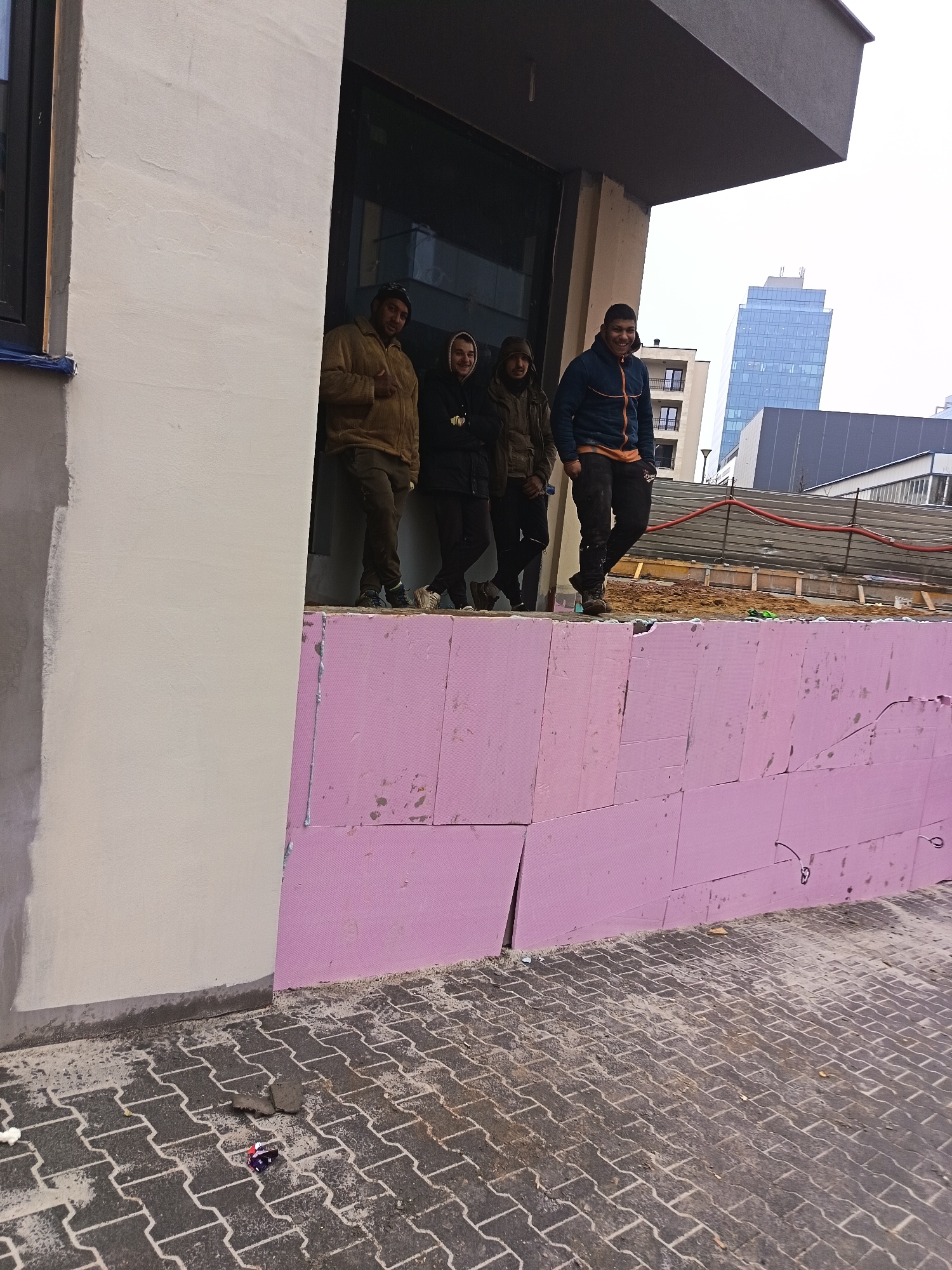 The image captures a scene in Sofia, Bulgaria, depicting four men standing on a partially constructed wall, likely at a construction site. The wall is made of pink insulation boards. The men are wearing casual clothing suitable for cool weather, with one in a brown jacket and hat, another in a hooded jacket, a third in a dark jacket, and the fourth in a blue and orange jacket and dark pants.
Behind the wall and the men, there is evidence of construction materials and dirt. Further back, a building under construction and other buildings, including a tall, modern skyscraper, are visible against a cloudy sky.
The foreground shows a paved area covered with some debris and dirt. The overall environment suggests an urban setting undergoing construction or renovation.