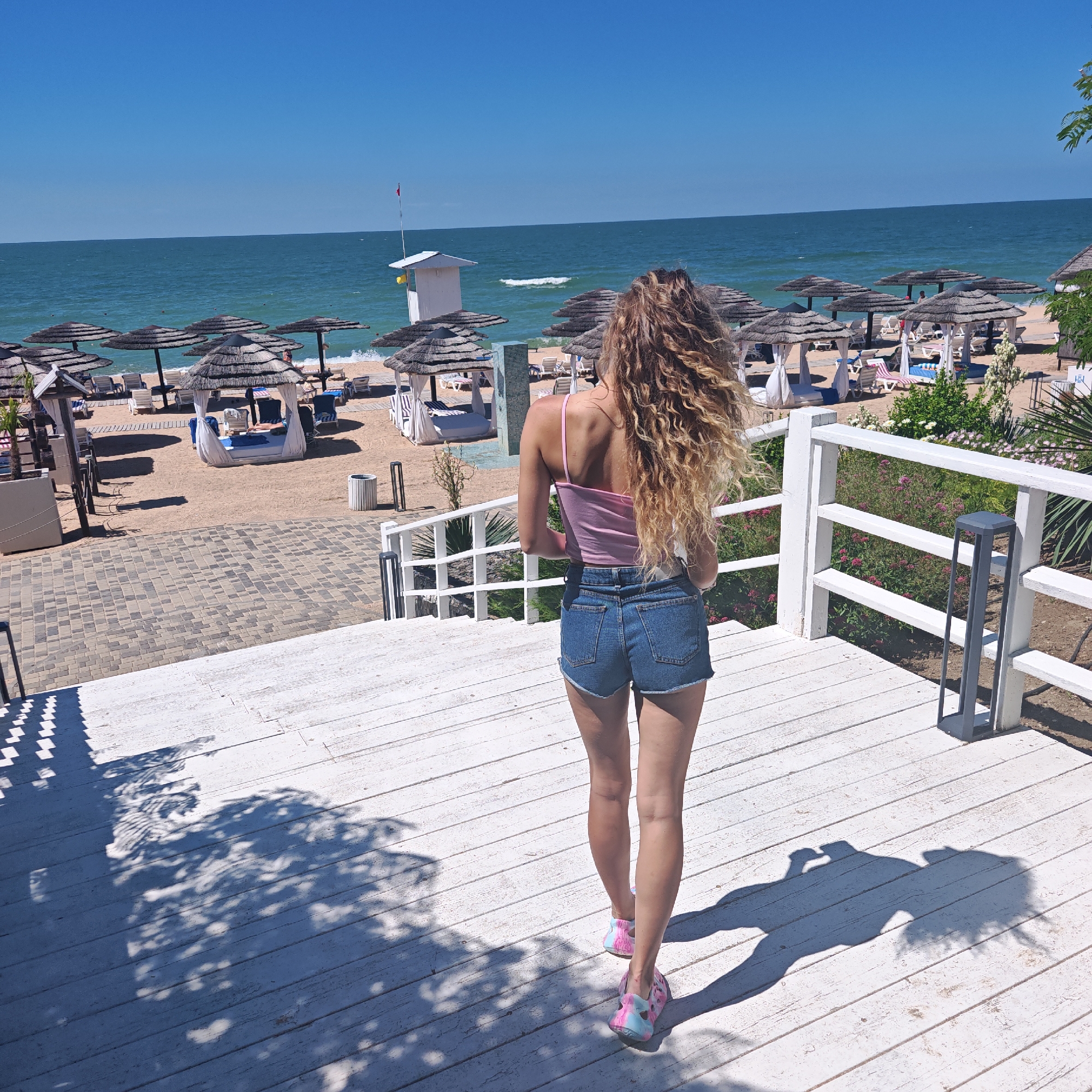 A woman with long, curly blonde hair is standing on a white wooden deck, facing away from the camera and towards a beach. She is wearing a pink tank top and denim shorts. The beach is lined with thatched-roof umbrellas and lounge chairs, and beyond that, the ocean stretches to the horizon under a clear blue sky. The scene appears to be a resort or a similar vacation spot.
