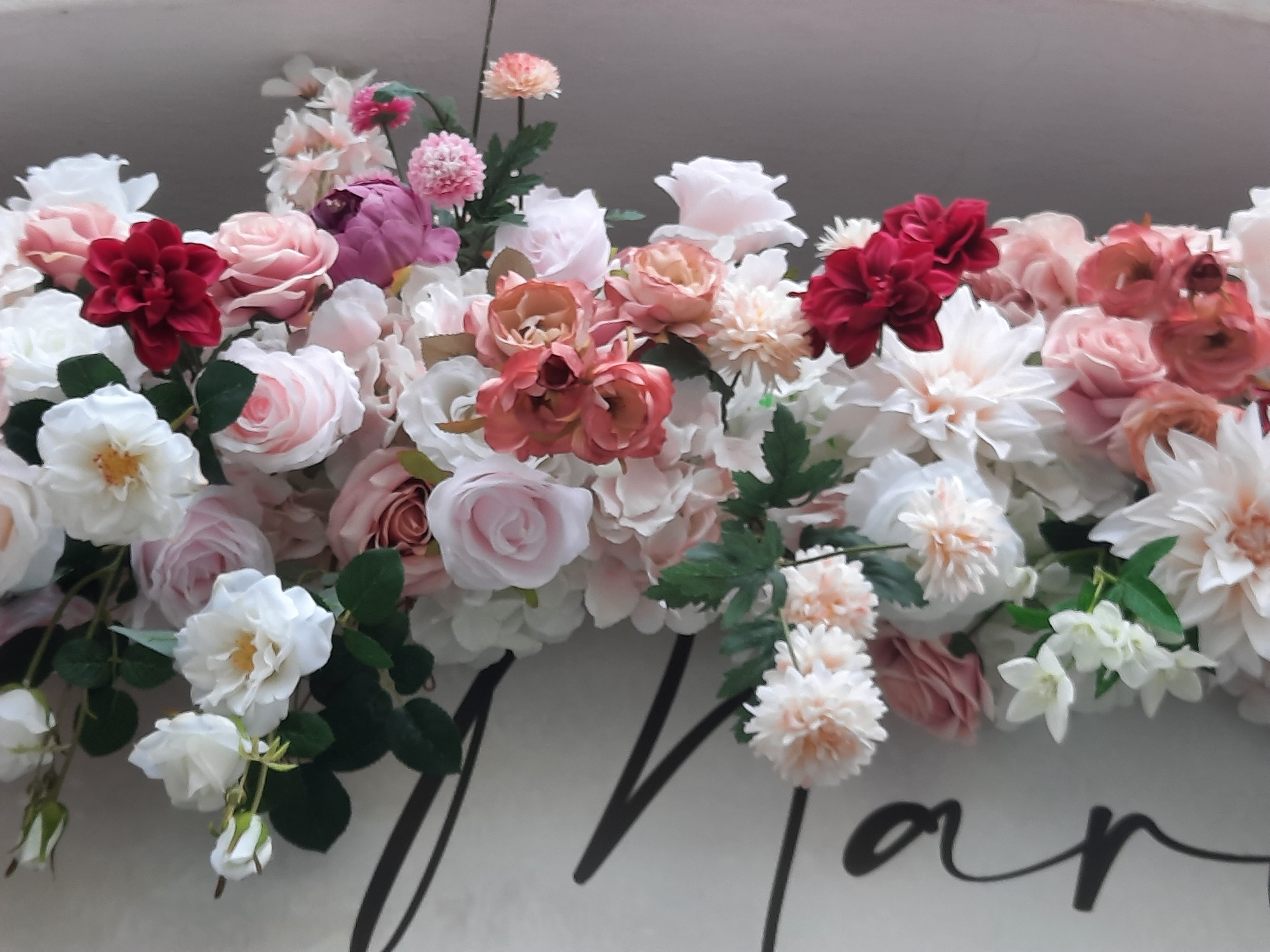 The image displays a close-up, eye-level shot of a floral arrangement that appears to be part of a larger decoration, possibly for an event. The arrangement consists of a variety of artificial flowers in shades of white, pink, peach, and deep red. Prominent flowers include large, multi-petaled roses, dahlias, and smaller, pom-pom-like blooms. Green leaves are interspersed throughout the arrangement. The flowers are densely packed, creating a lush and textured visual.
Beneath the floral arrangement, a portion of a white surface is visible, with a large, dark, cursive font forming letters. The visible letters appear to be part of the word "Van" followed by other indistinct characters. This suggests the floral arrangement is likely placed above or in front of lettering on a sign or backdrop. The background is a neutral, muted gray, with a slight gradient. There are no people or animals present in the image. The overall impression is one of decorative floral artistry.
Without further information or context, it is impossible to determine the specific location in the city of Rosenheim, Germany, where this image was taken. The scene itself provides no geographical indicators specific to Rosenheim.