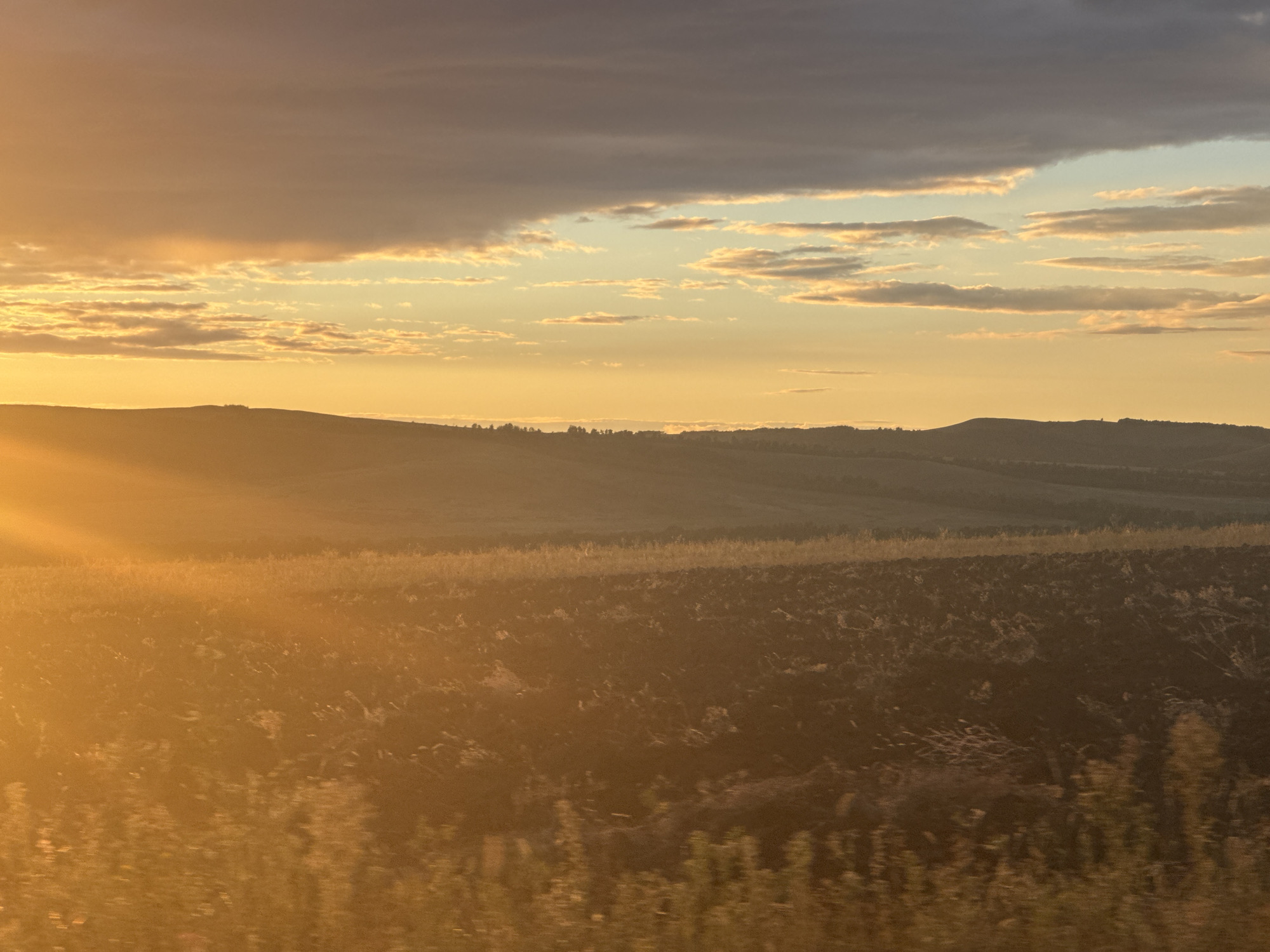 The image depicts a rural landscape at sunset, featuring rolling hills and a cultivated field. The sky is filled with dramatic clouds, ranging from dark gray to bright orange and yellow, illuminated by the setting sun. The sun's rays cast a warm glow across the scene, creating a strong contrast between light and shadow. The foreground shows a dark, tilled field with visible patterns of soil. In the middle ground, the terrain is gentler, with faint vegetation and a distant tree line. The background consists of the silhouette of hills against the colorful sky. The location is identified as Vasil'evka, Russia. There are no people or animals visible in the image. The overall impression is one of natural beauty and the serene end of a day.